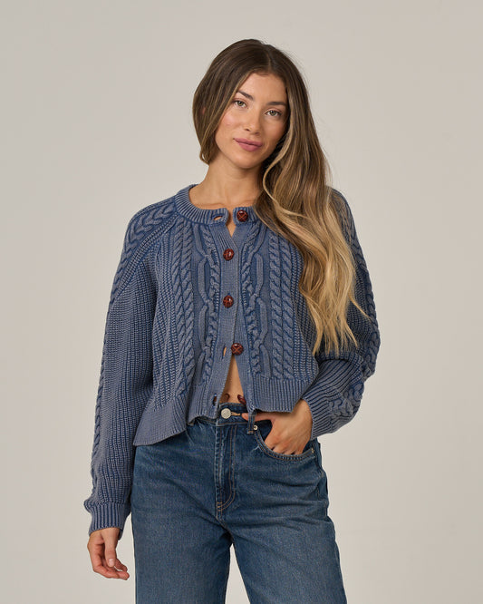 A woman with long, wavy hair wears the Salty Crew Fisherman Cardigan in Flint—crafted from soft cotton sweater material with brown buttons—paired with blue jeans, standing against a plain light background.
