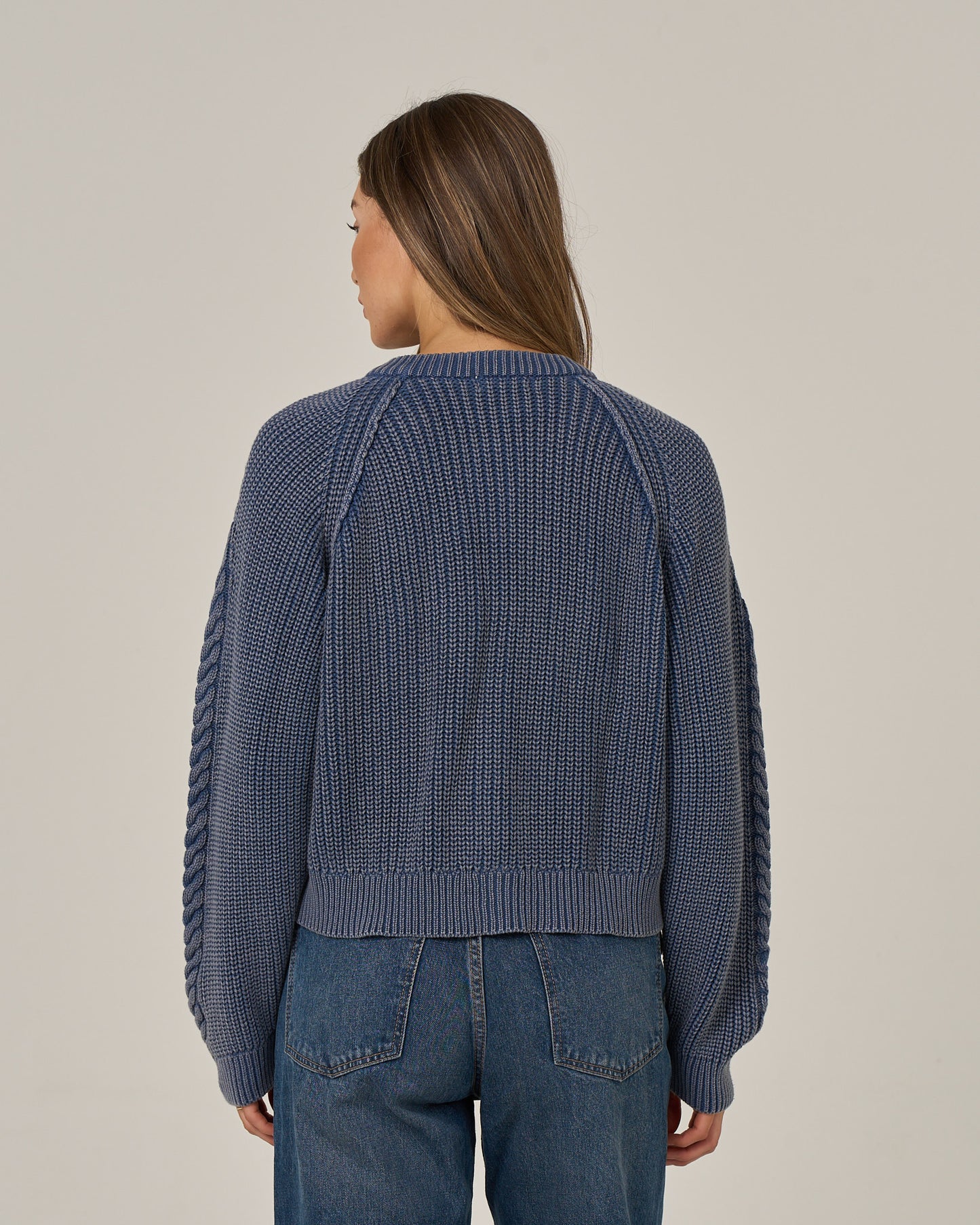 A woman with long brown hair is standing facing away, wearing the Salty Crew Fisherman Cardigan - Flint and vintage wash blue jeans, against a plain light background.