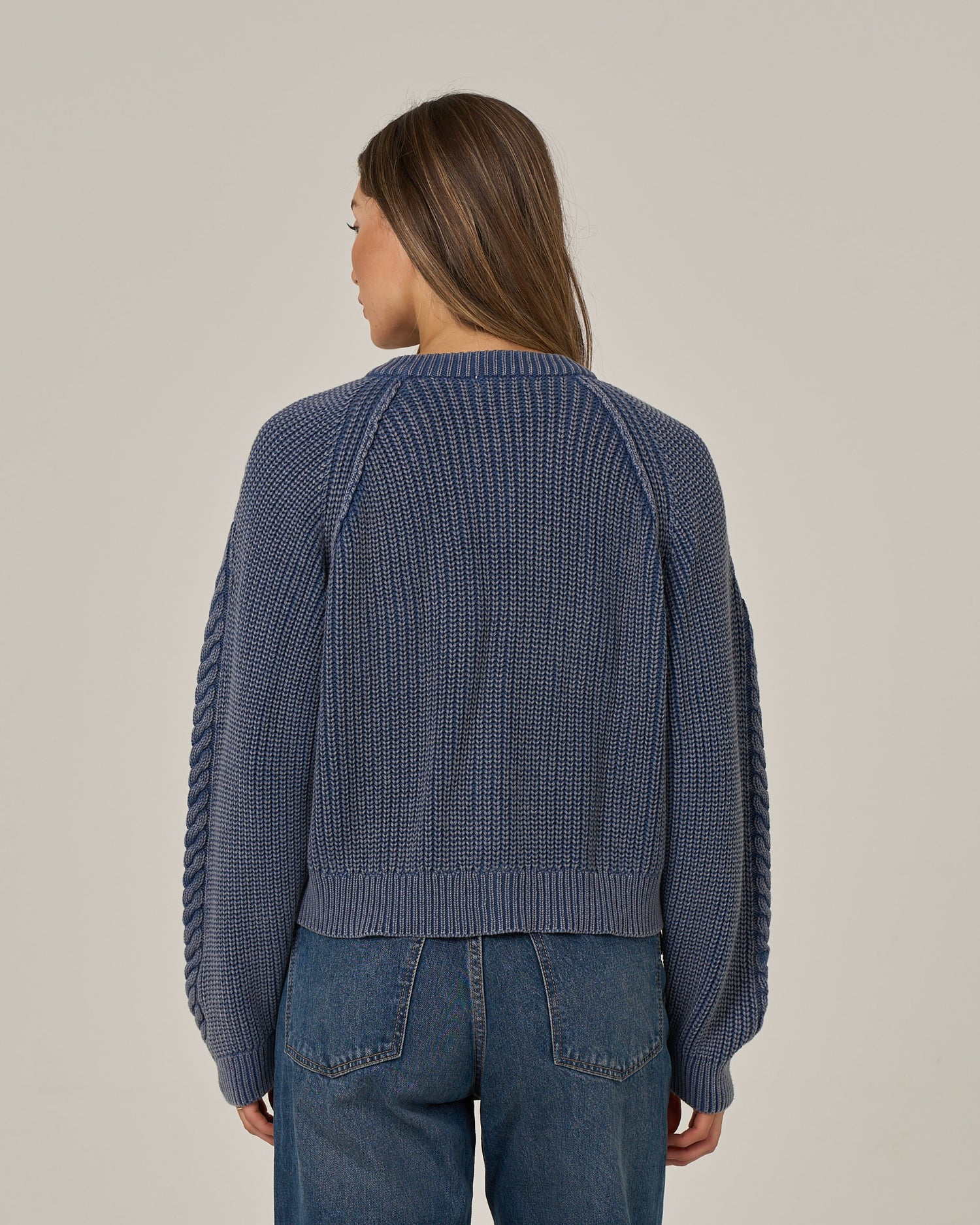 A woman with long brown hair is standing facing away, wearing the Salty Crew Fisherman Cardigan - Flint and vintage wash blue jeans, against a plain light background.