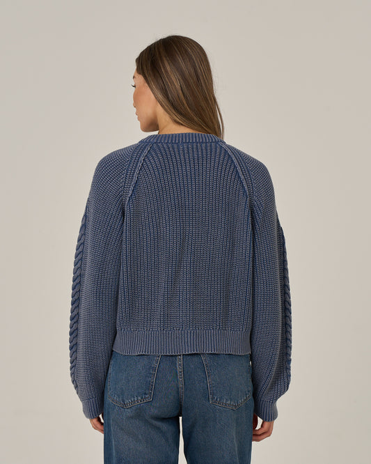 A woman with long brown hair is standing facing away, wearing the Salty Crew Fisherman Cardigan - Flint and vintage wash blue jeans, against a plain light background.