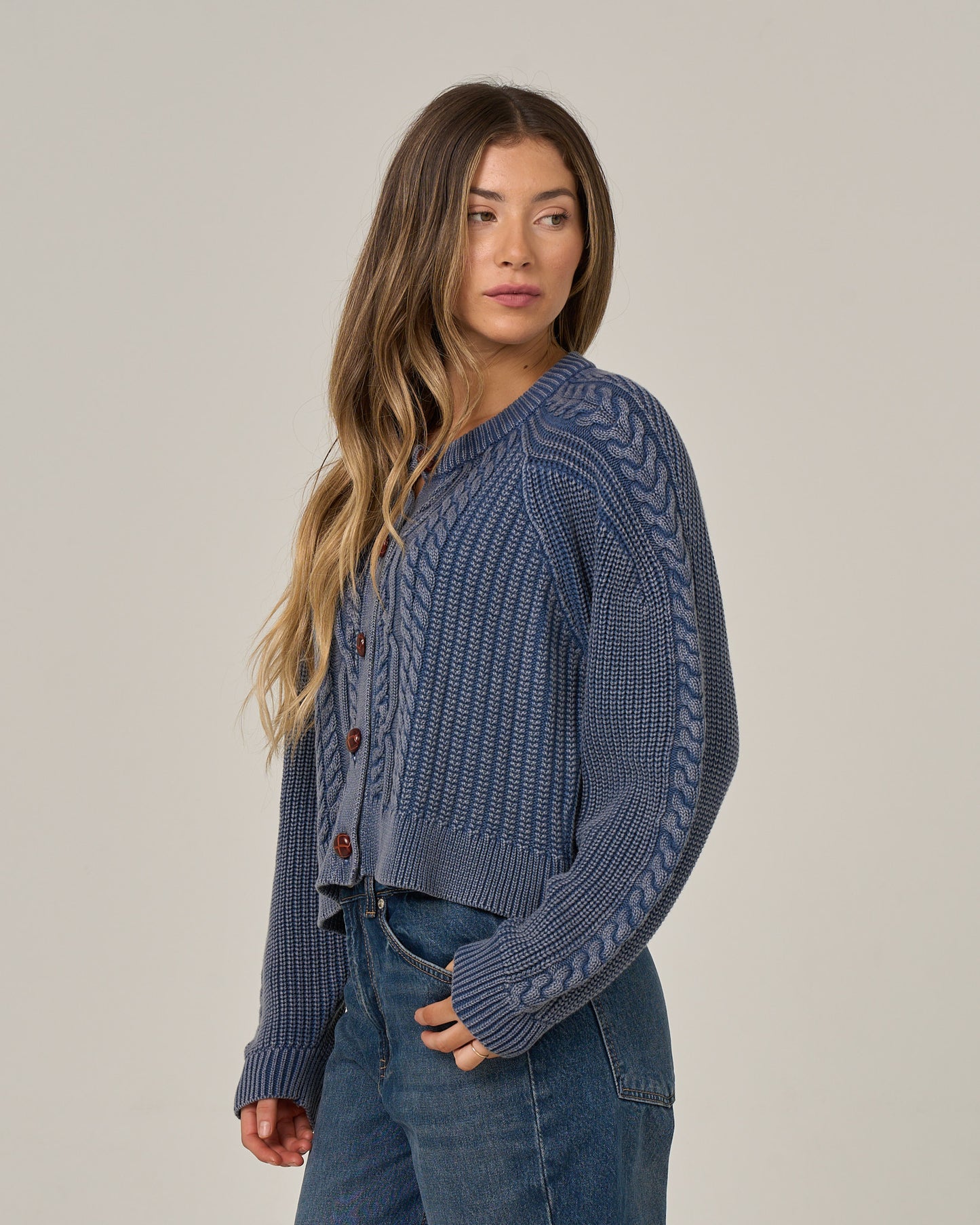 A woman with long wavy hair wears the Salty Crew Fisherman Cardigan in Flint and blue jeans, standing against a plain light background, looking to her left with a relaxed expression.