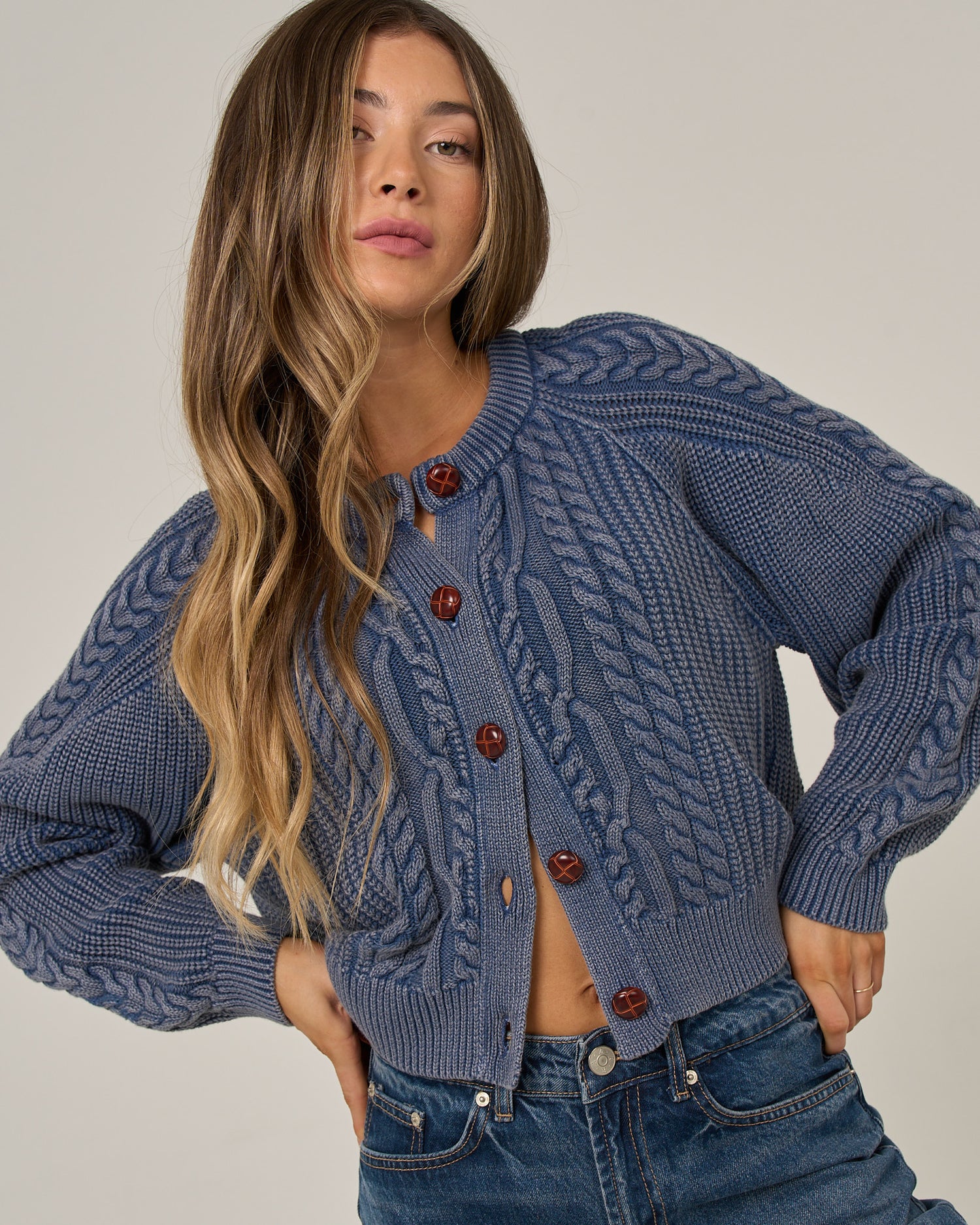 A woman with long wavy hair wears the Salty Crew Fisherman Cardigan in Flint—a blue cotton cable-knit sweater with brown buttons—paired with vintage-wash blue jeans, posing with one hand on her hip against a neutral background.