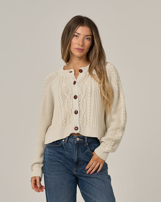 A woman wearing the Salty Crew Fisherman Cardigan in Stone and vintage wash blue jeans stands against a plain light background, looking at the camera with a neutral expression.