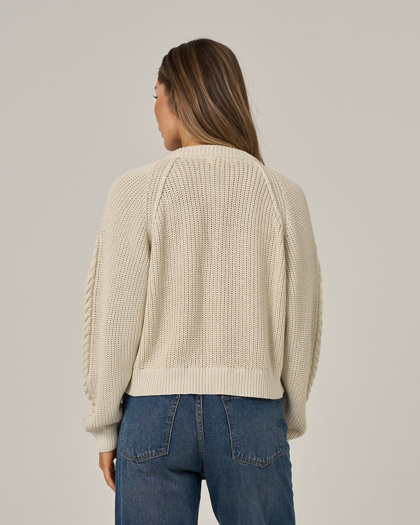 A woman with long brown hair, wearing the Salty Crew Fisherman Cardigan in Stone and vintage-wash blue jeans, stands facing away from the camera against a plain light background.
