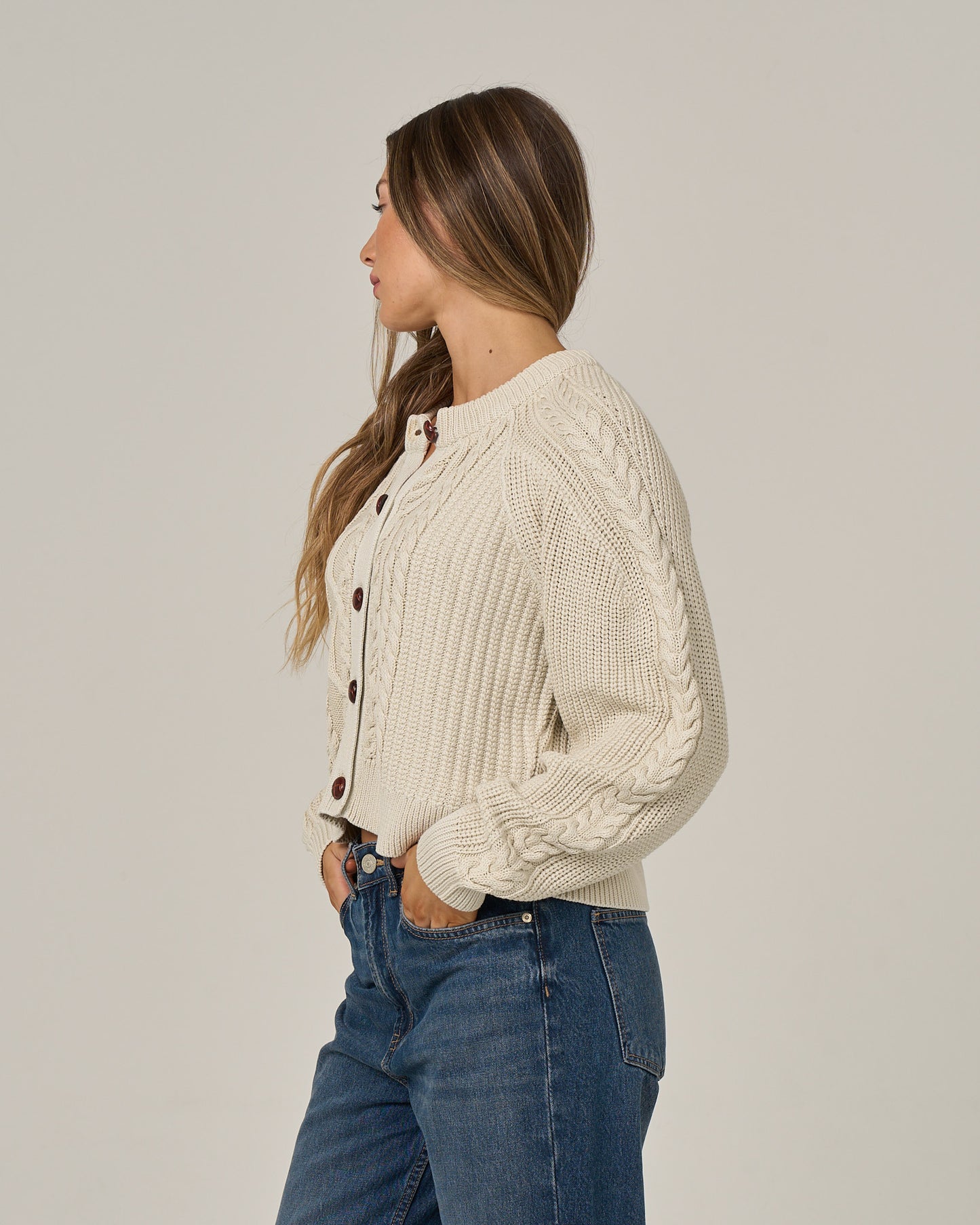 A woman stands in profile against a plain light background, wearing the Salty Crew Fisherman Cardigan in Stone, featuring brown buttons and cable knit detailing, paired with vintage wash blue jeans and her hands in her pockets as she looks to the left.