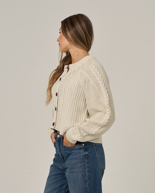 A woman stands in profile against a plain light background, wearing the Salty Crew Fisherman Cardigan in Stone, featuring brown buttons and cable knit detailing, paired with vintage wash blue jeans and her hands in her pockets as she looks to the left.
