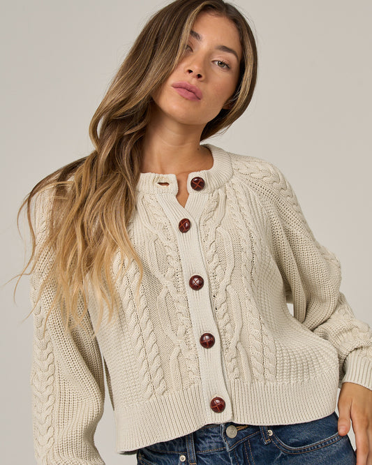 A woman with long, wavy hair wears the Salty Crew Fisherman Cardigan in Stone, featuring large brown buttons, paired with vintage-wash blue jeans as she stands against a plain background.