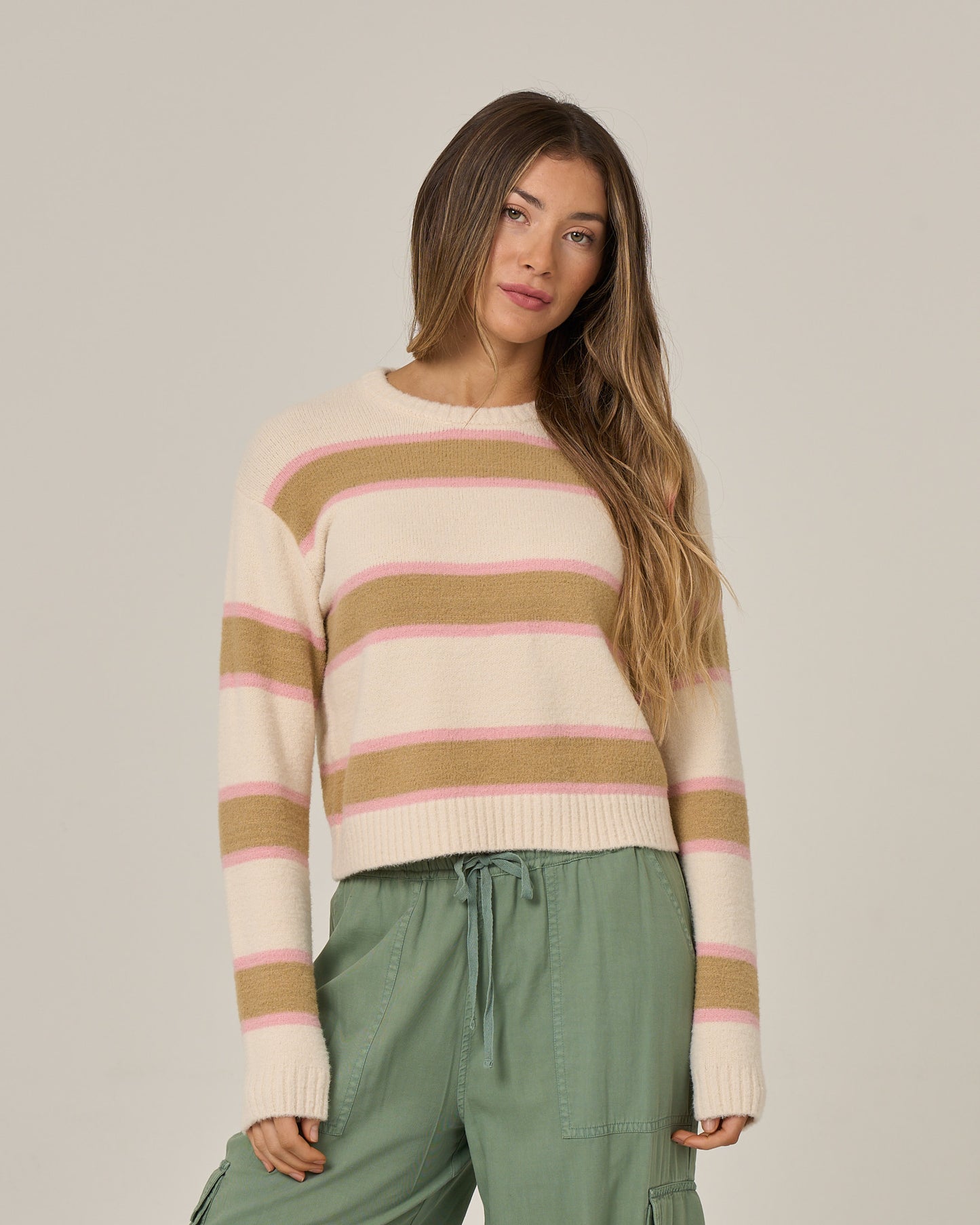 A woman with long hair wears the East Cape Sweater in Birch by Salty Crew—a fuzzy, stripe crew neck sweater in cream with pink and tan stripes—paired with green cargo pants, standing against a plain light background.