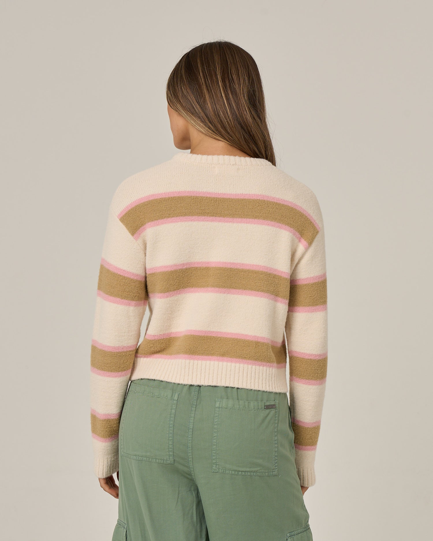 A person with long brown hair wears the Salty Crew East Cape Sweater in Birch, featuring fuzzy cream fabric with pink and tan stripes, paired with green cargo pants, facing away from the camera against a plain background.