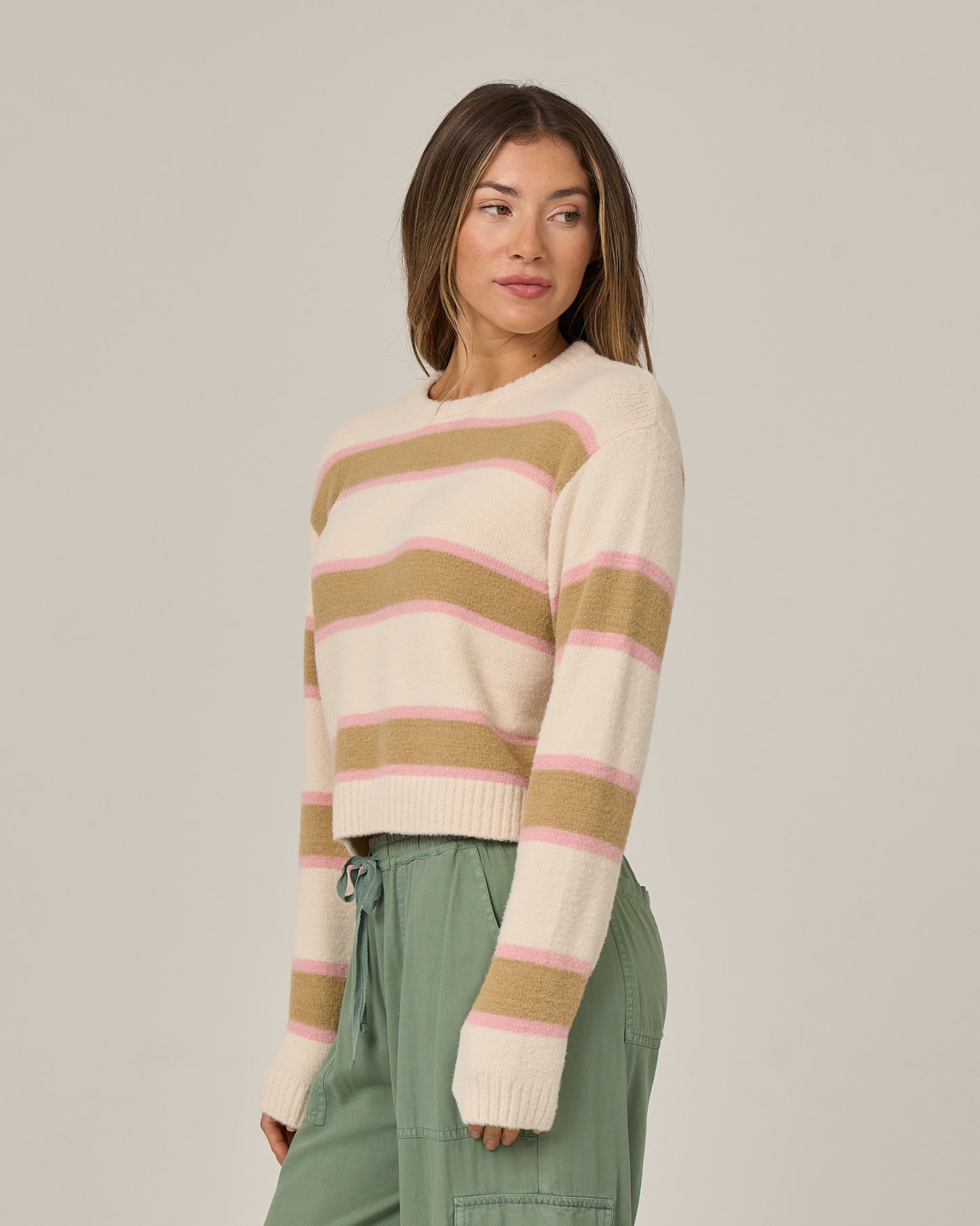 A woman wearing the Salty Crew East Cape Sweater in Birch—a fuzzy sweater with tan and pink horizontal stripes over a cream base—paired with green pants, stands against a plain background, looking to the side.