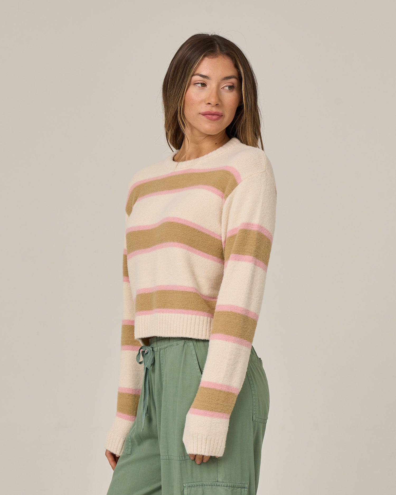 A woman wearing the Salty Crew East Cape Sweater in Birch—a fuzzy sweater with tan and pink horizontal stripes over a cream base—paired with green pants, stands against a plain background, looking to the side.