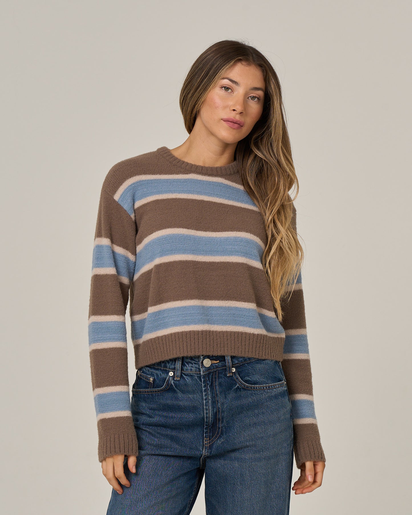 A woman stands facing forward, wearing the Salty Crew East Cape Sweater in Washed Brown with blue jeans, against a plain light background.