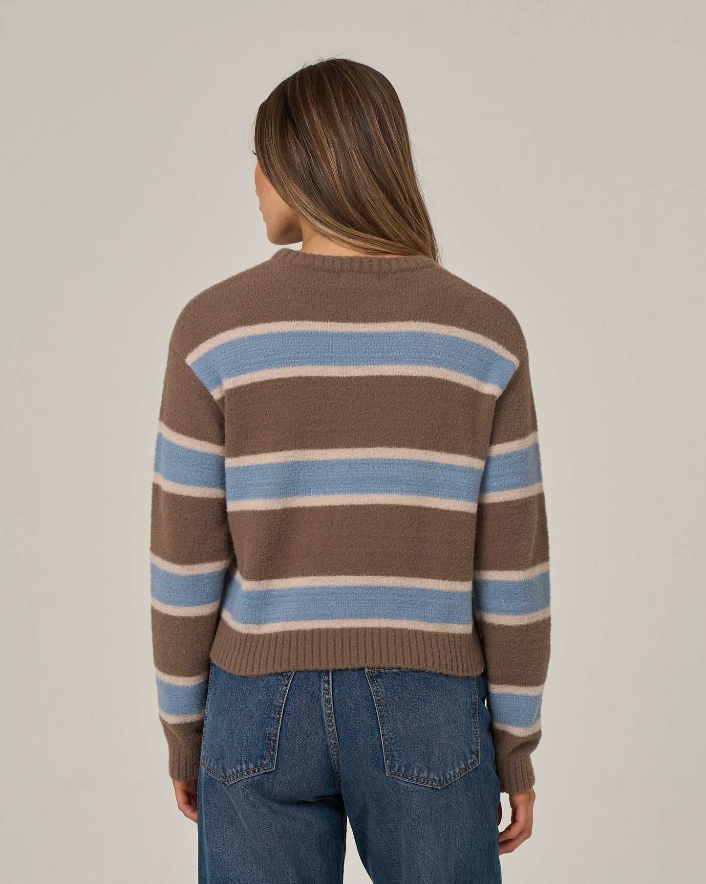 A person with long brown hair, wearing the Salty Crew East Cape Sweater in Washed Brown and blue jeans, stands with their back to the camera against a plain light background.