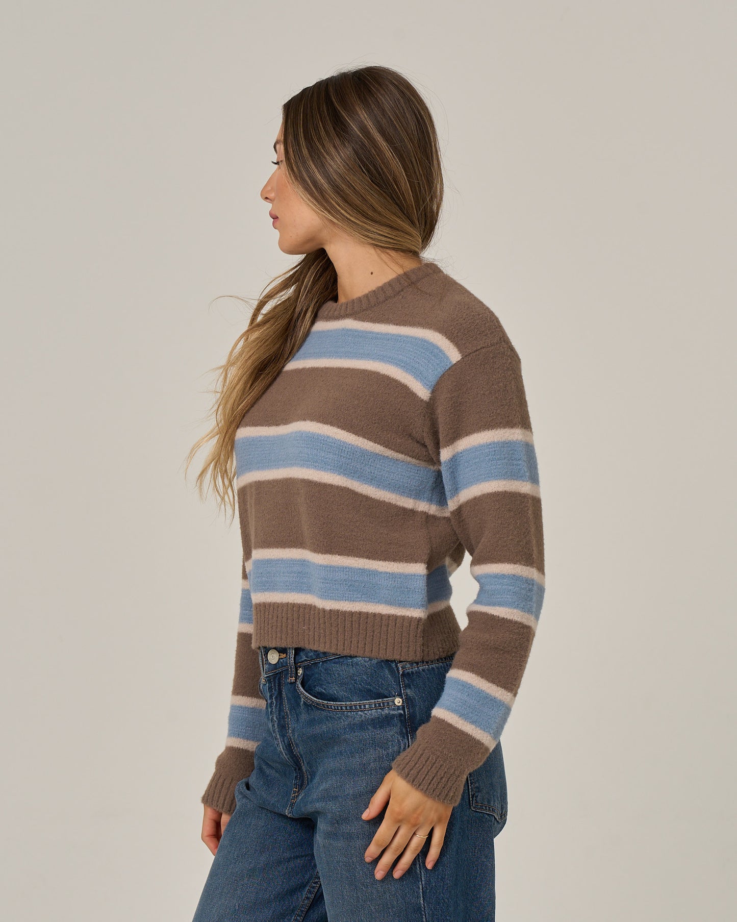 A woman with long brown hair is wearing the Salty Crew East Cape Sweater in washed brown, featuring blue and white stripes, paired with blue jeans while standing sideways against a plain background.