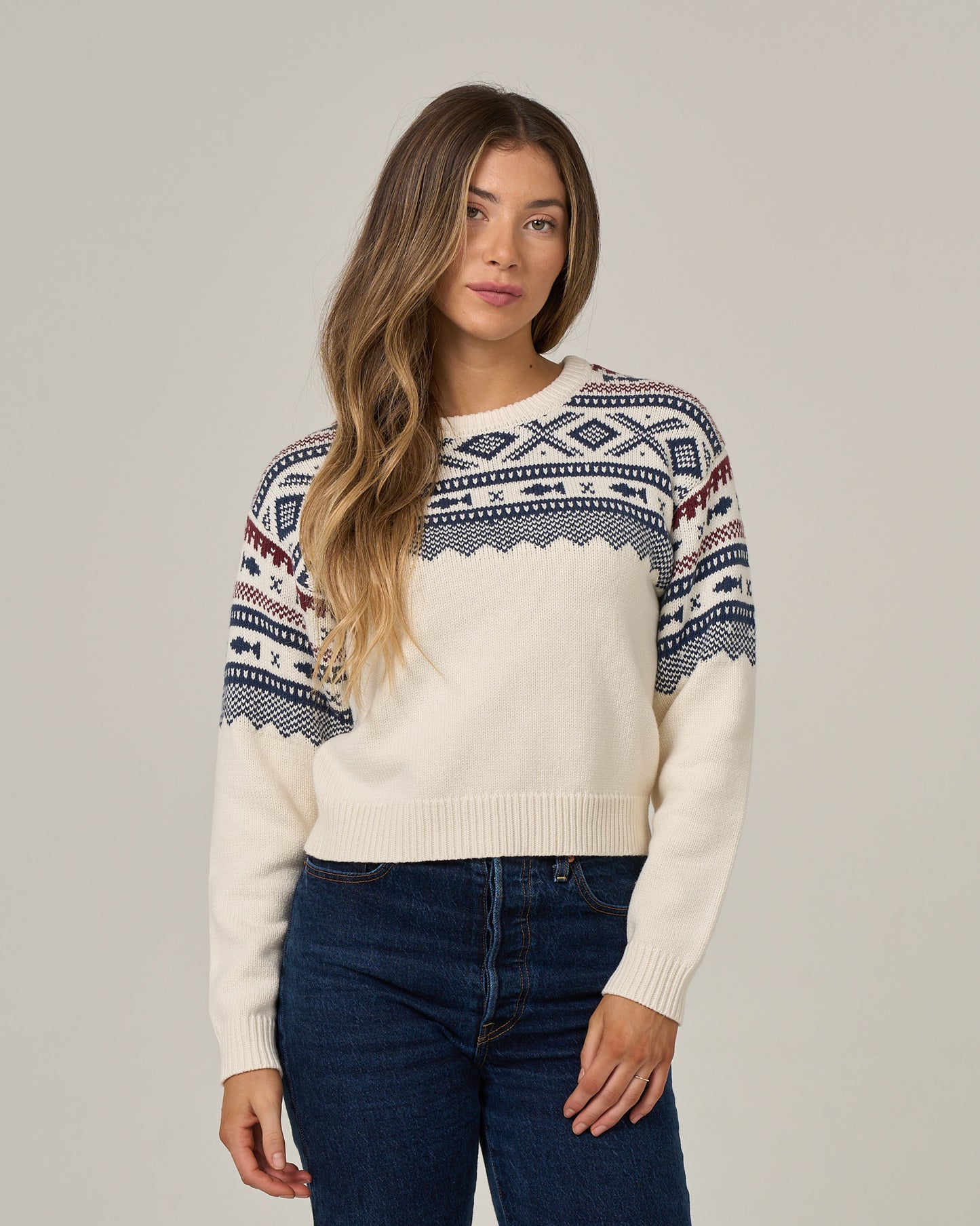 A woman with long, wavy hair wears the Salty Crew Harbor Sweater in Off White, featuring a blue and red jacquard pattern, paired with dark blue jeans as she stands against a plain light background.