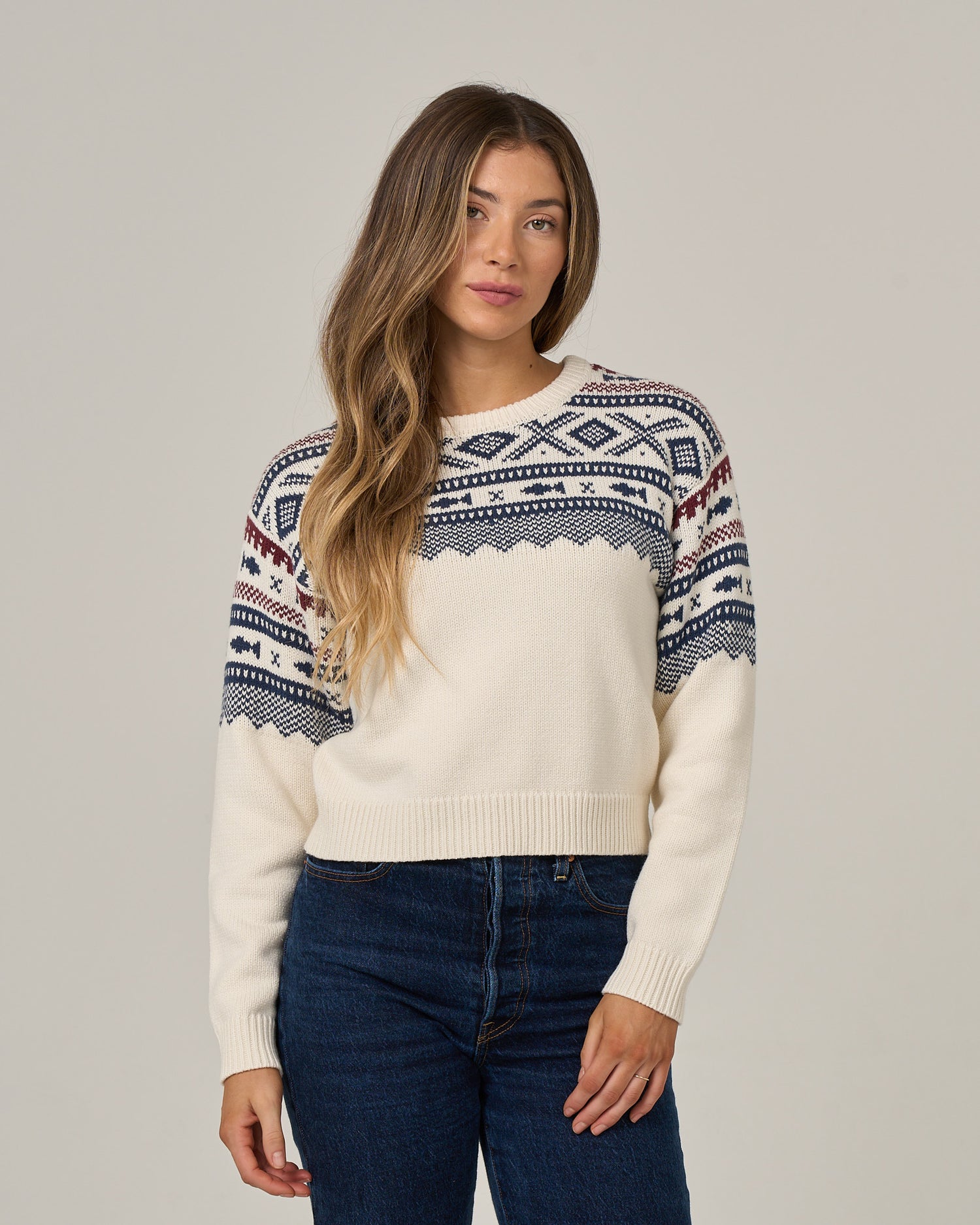 A woman with long, wavy hair wears the Salty Crew Harbor Sweater in Off White, featuring a blue and red jacquard pattern, paired with dark blue jeans as she stands against a plain light background.