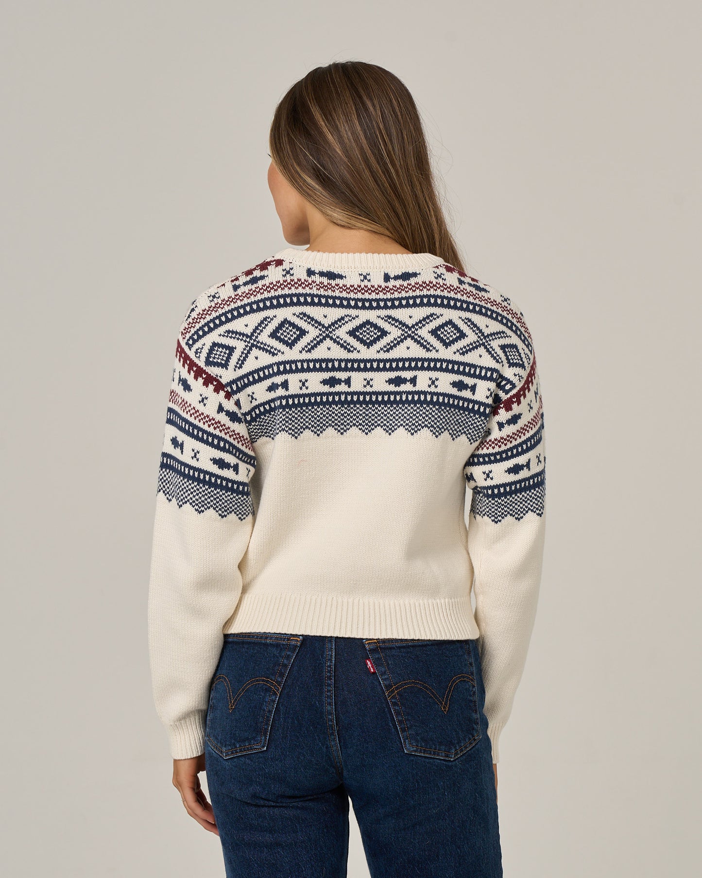 A person with long hair, seen from behind against a plain gray background, is wearing the Salty Crew Harbor Sweater in Off White—featuring blue and red jacquard patterns—and blue jeans.