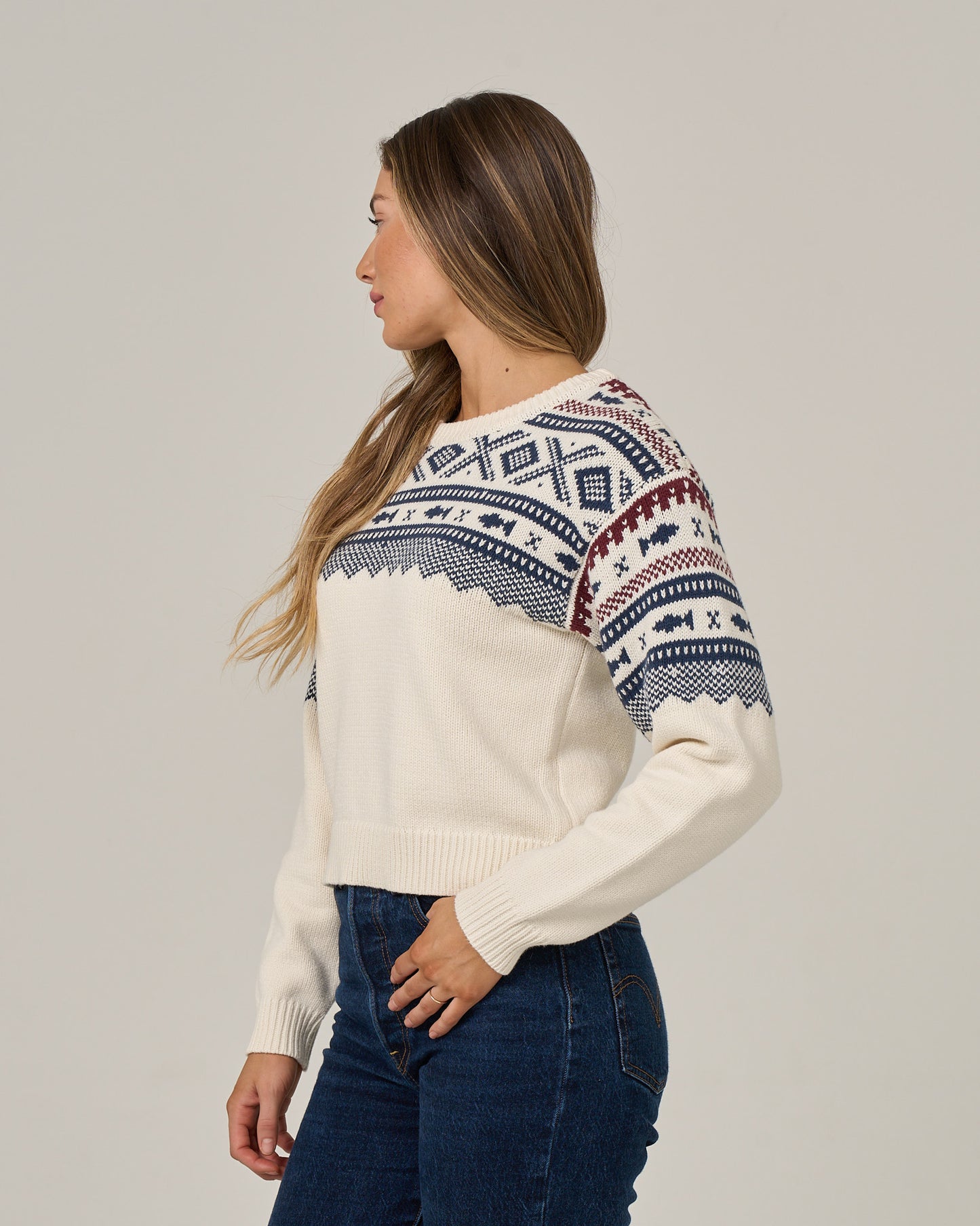 A woman with long, straight brown hair stands sideways against a plain light background, looking to the left. She is wearing Salty Crew’s Harbor Sweater in Off White, featuring blue and red jacquard patterns and a crew neckline, paired with dark blue jeans.