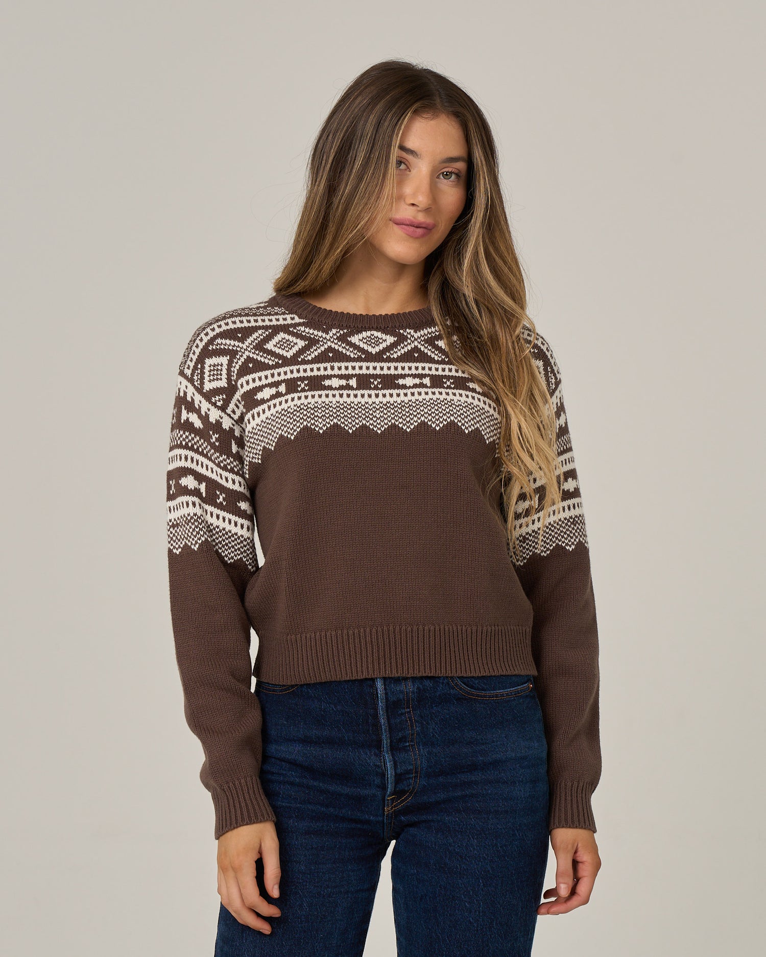 A woman with long hair wears the Salty Crew Harbor Sweater in washed brown, featuring white jacquard patterns, paired with blue jeans and standing against a plain light background.