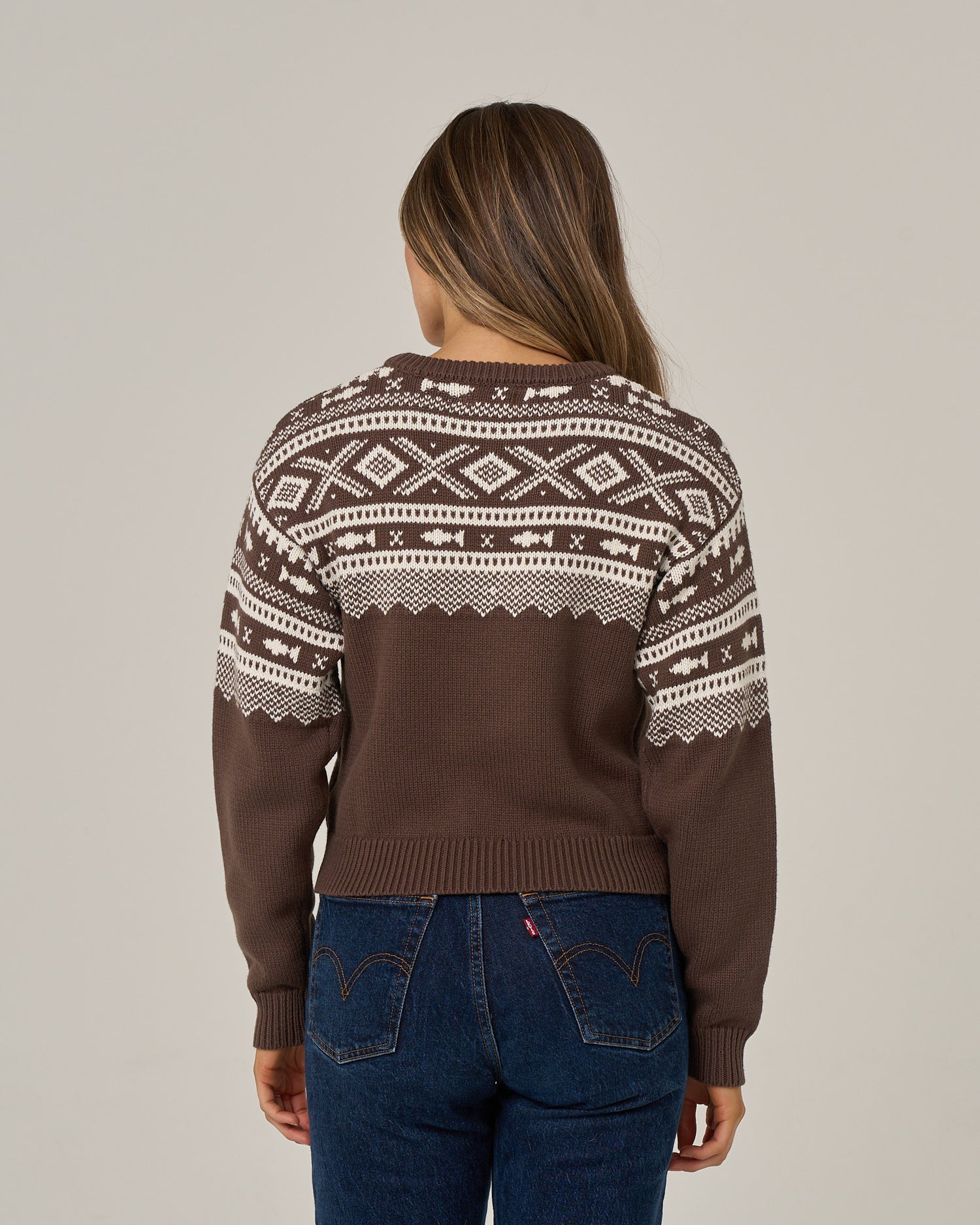 A person with long brown hair stands with their back to the camera against a plain light background, wearing the Salty Crew Harbor Sweater in Washed Brown—a cotton crew neck sweater featuring a brown and white jacquard pattern—paired with blue jeans.