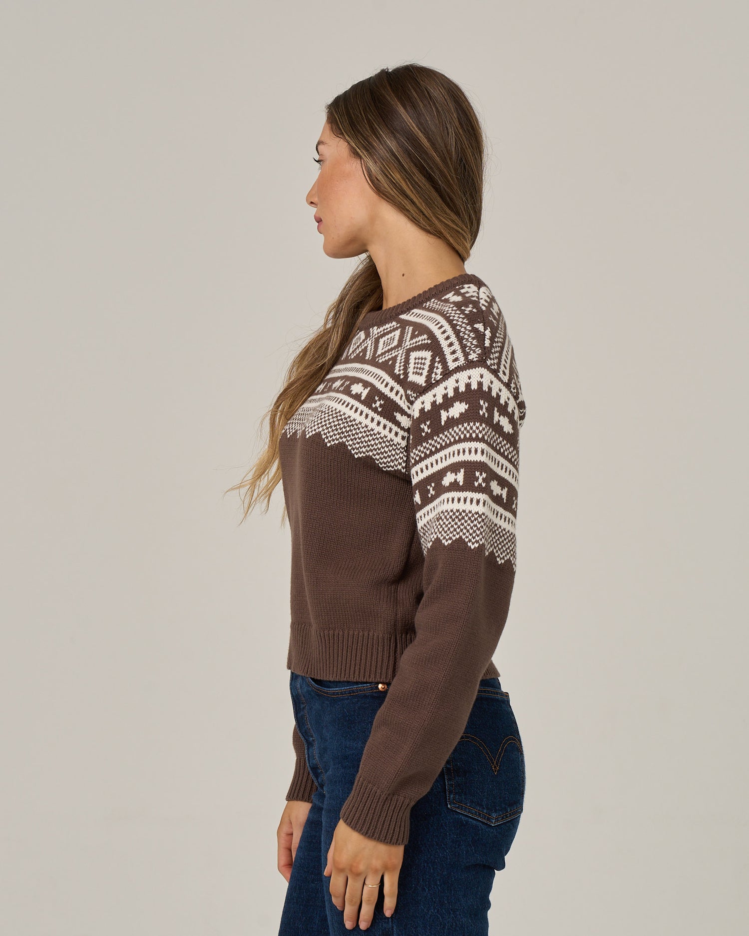A woman in profile is wearing the Salty Crew Harbor Sweater in washed brown—a brown and white jacquard pattern crew neck—paired with blue jeans, standing against a plain light background.