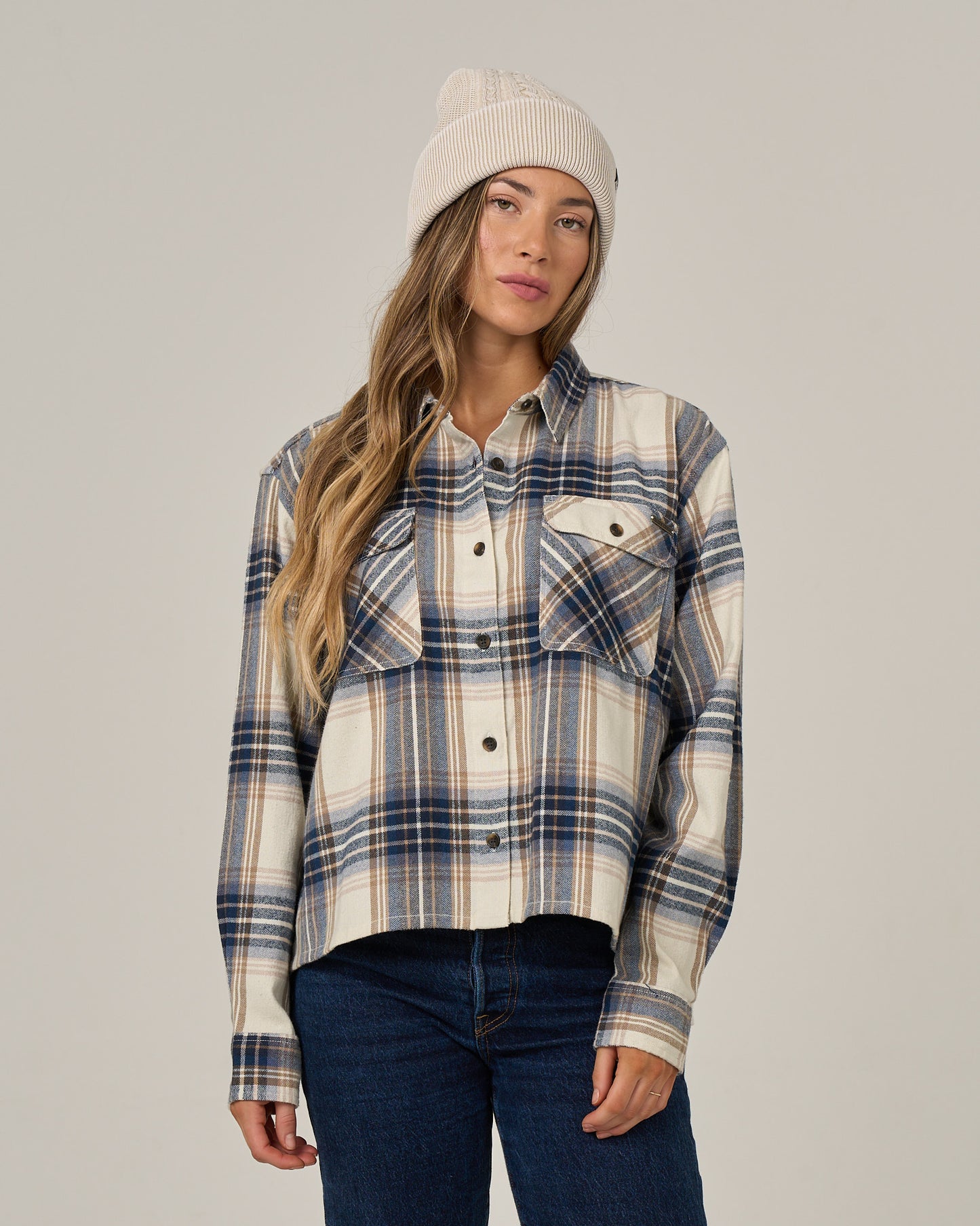 A woman wearing the Salty Crew Nomad Flannel in Birch and a cream beanie stands against a plain background with a neutral expression.