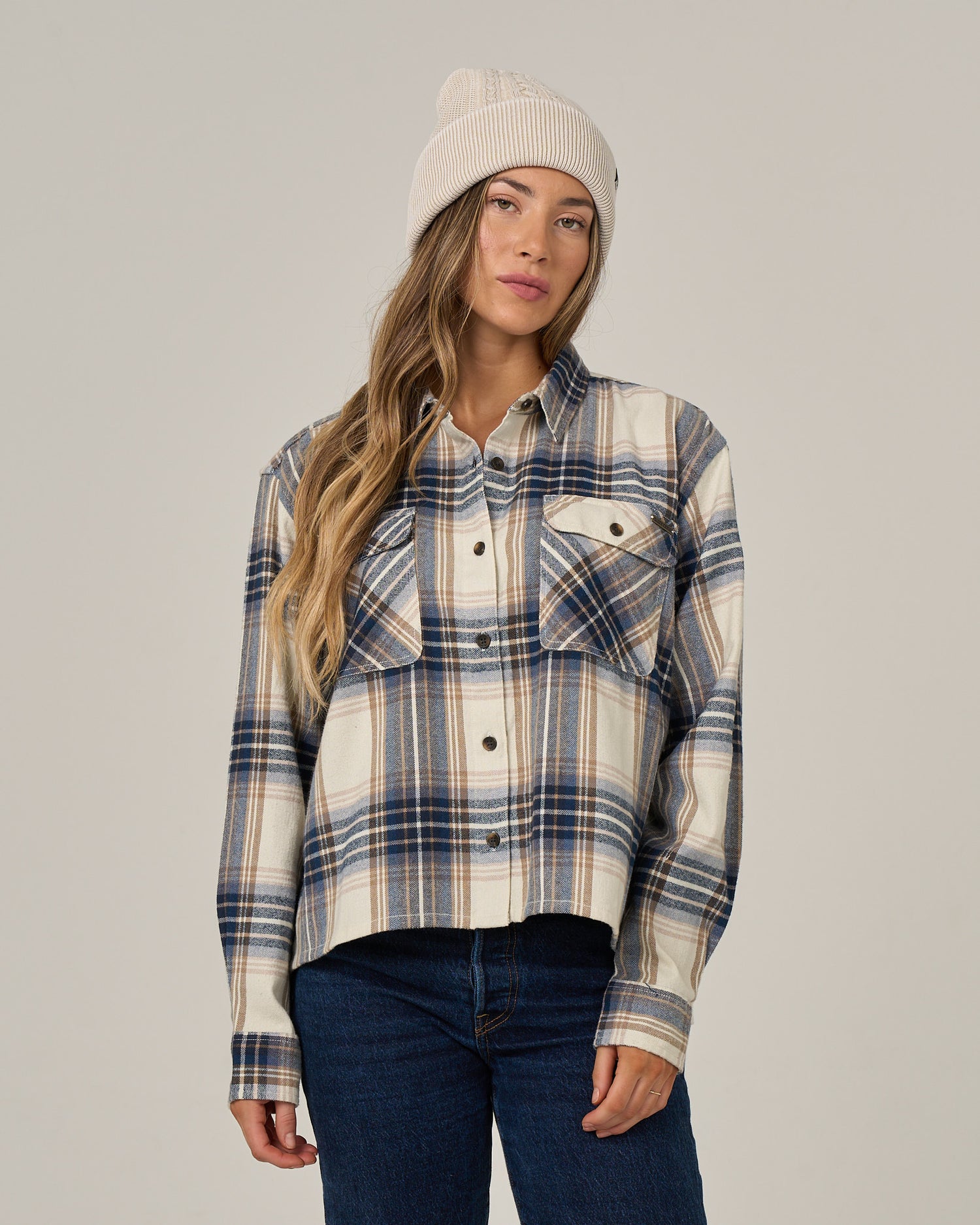 A woman wearing the Salty Crew Nomad Flannel in Birch and a cream beanie stands against a plain background with a neutral expression.