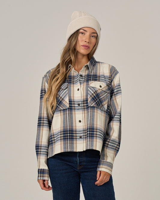 A woman wearing the Salty Crew Nomad Flannel in Birch and a cream beanie stands against a plain background with a neutral expression.