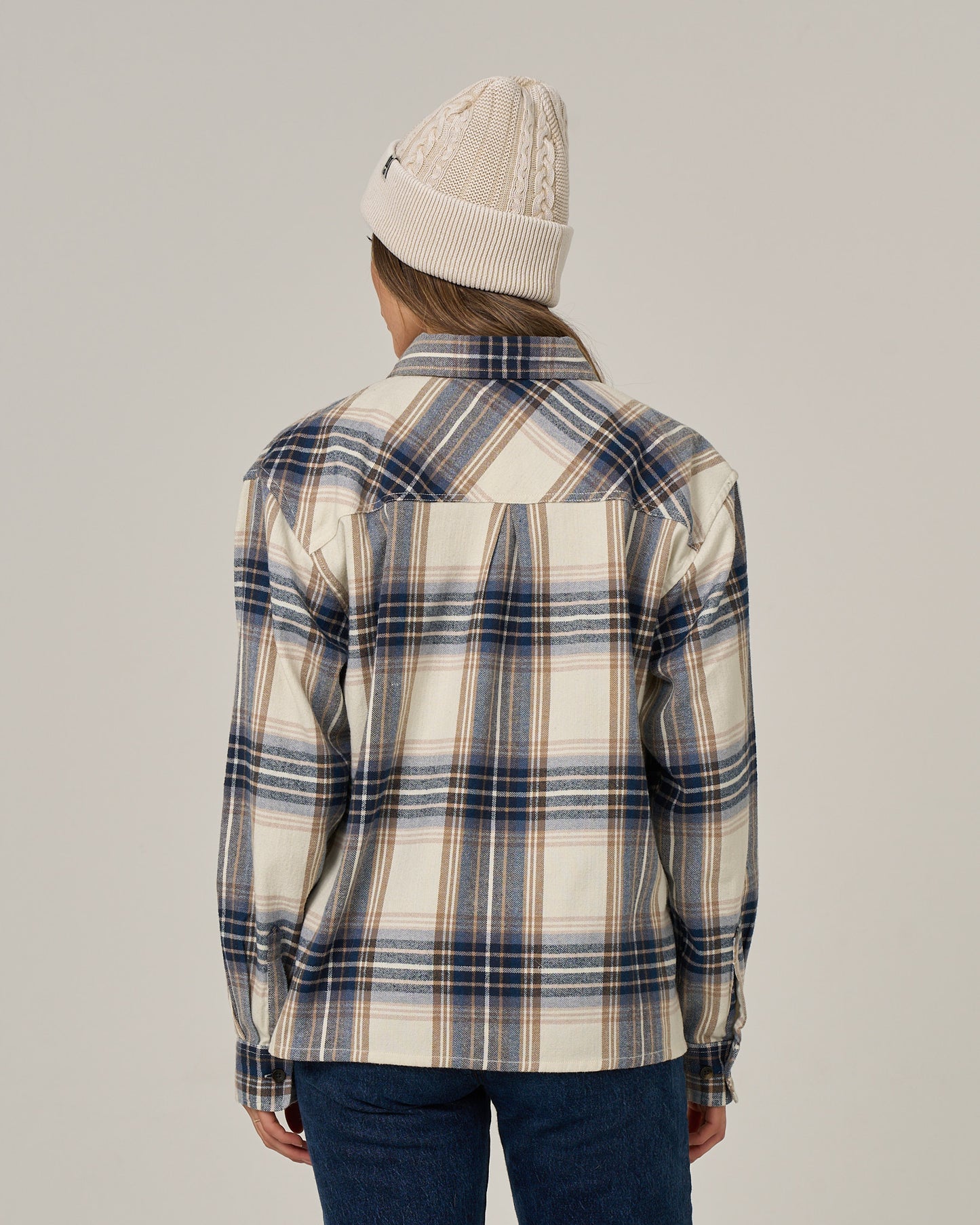 A person wearing a cream knit beanie and the Salty Crew Nomad Flannel - Birch, an oversized blue, white, and beige cotton polyester button-down shirt, stands facing away from the camera against a plain background.