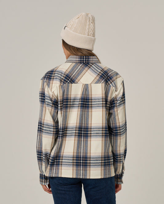 A person wearing a cream knit beanie and the Salty Crew Nomad Flannel - Birch, an oversized blue, white, and beige cotton polyester button-down shirt, stands facing away from the camera against a plain background.