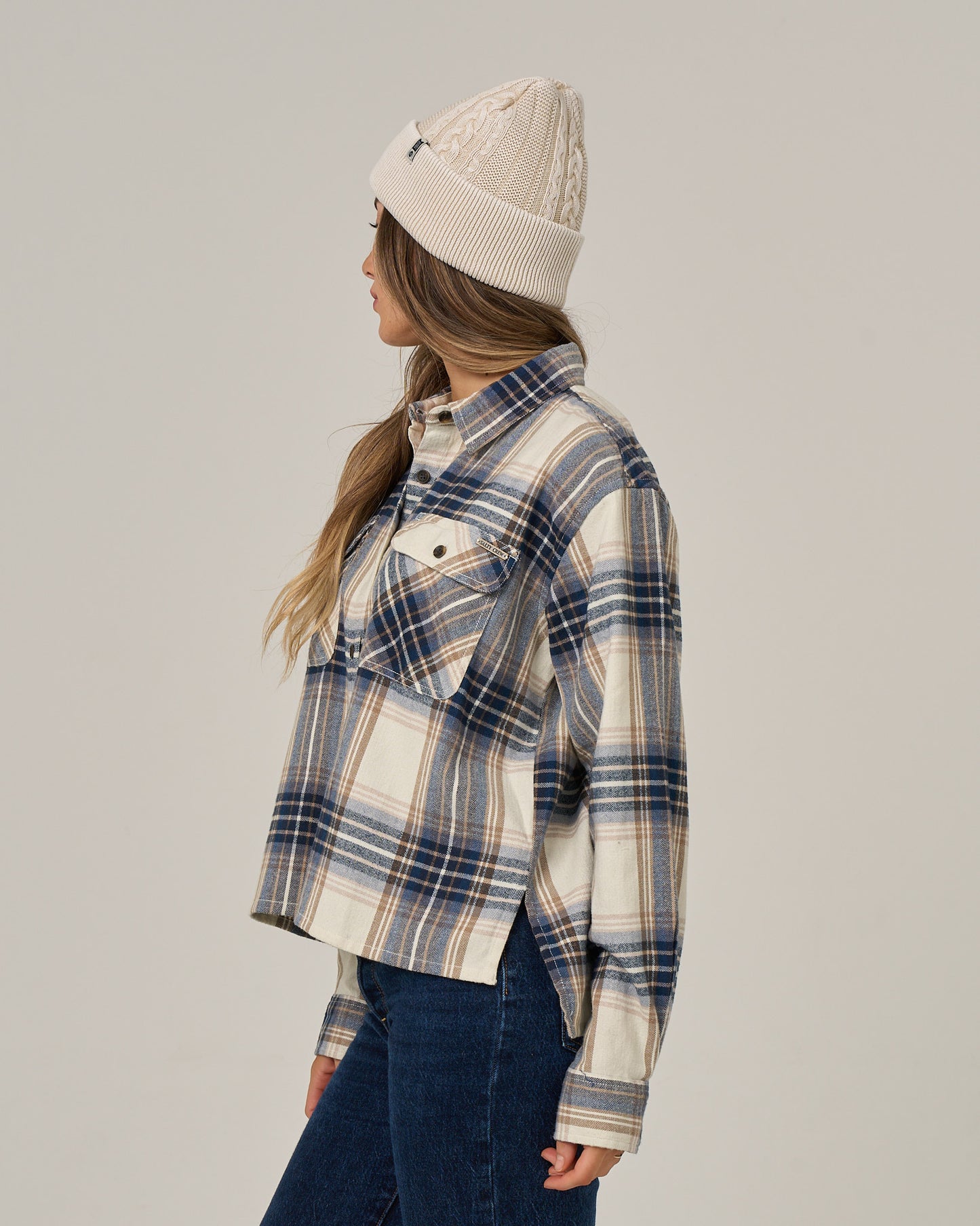 A person wearing a cream knit beanie, blue jeans, and the Salty Crew “Nomad Flannel - Birch” button-down shirt stands facing left against a plain light background.