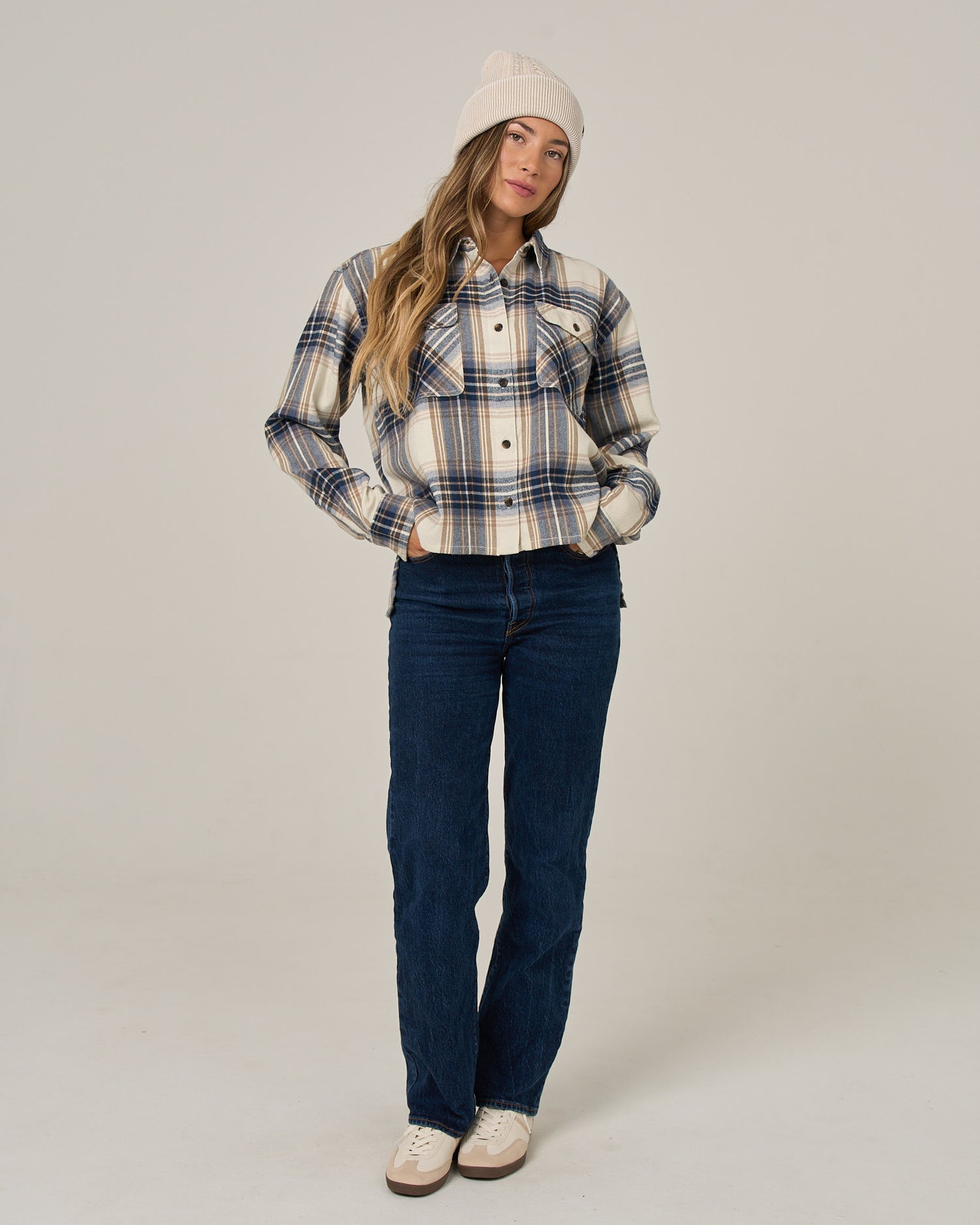 A woman in the Salty Crew Nomad Flannel - Birch, blue jeans, and a beige beanie stands with her hands in her pockets against a plain background.
