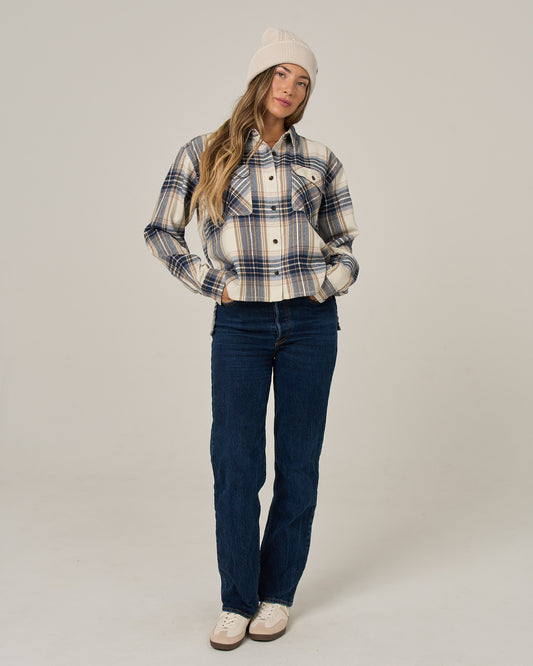 A woman in the Salty Crew Nomad Flannel - Birch, blue jeans, and a beige beanie stands with her hands in her pockets against a plain background.