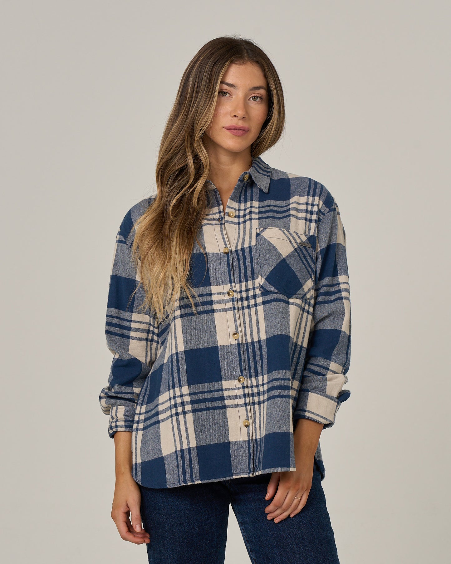 A woman with long wavy hair wears the Salty Crew Out West Flannel - Navy, a blue and beige western style plaid button-up shirt crafted from soft cotton flannel, paired with dark jeans, standing against a plain light background.