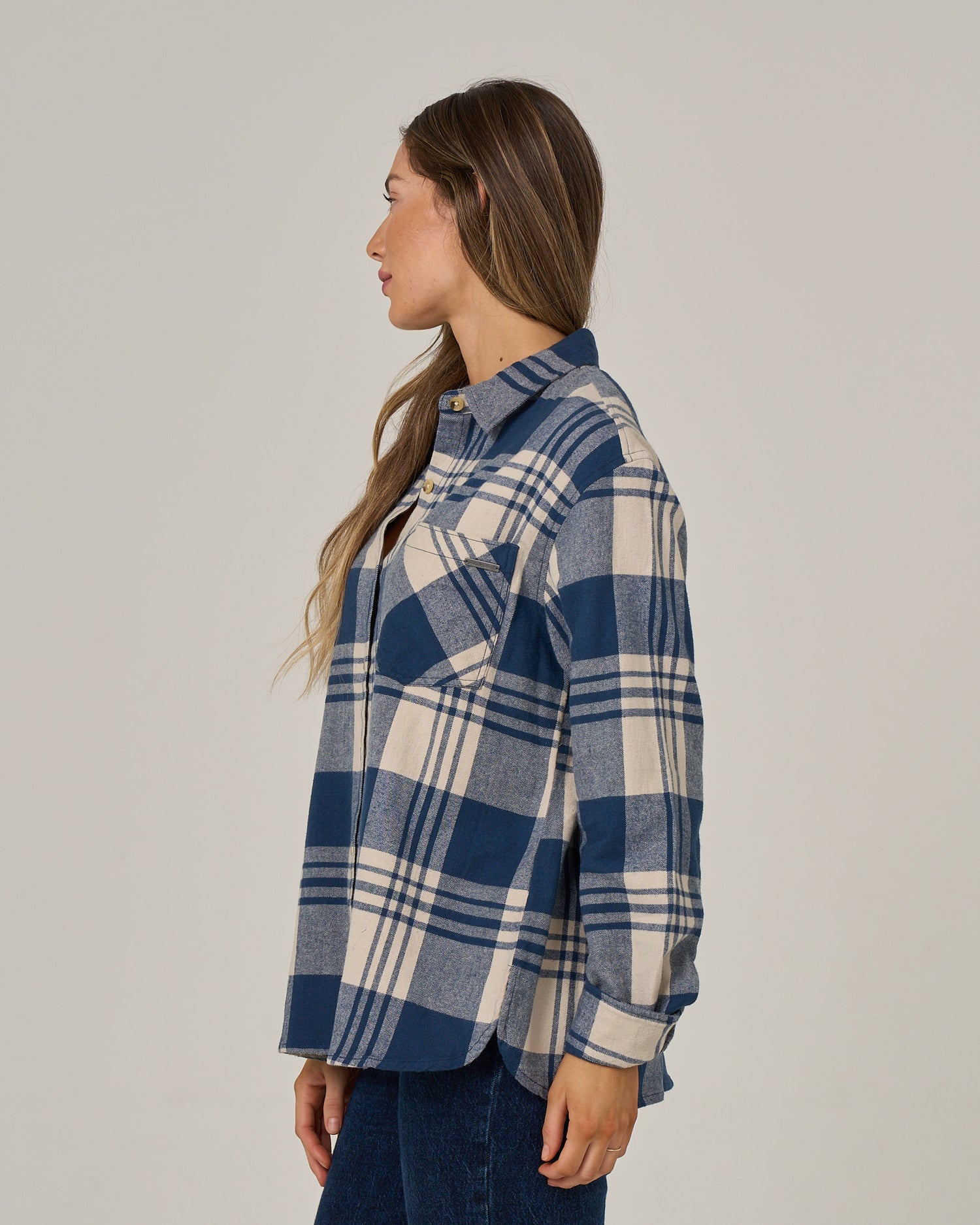 A woman with long brown hair stands sideways against a plain light background, wearing the Salty Crew Out West Flannel in Navy paired with dark jeans.