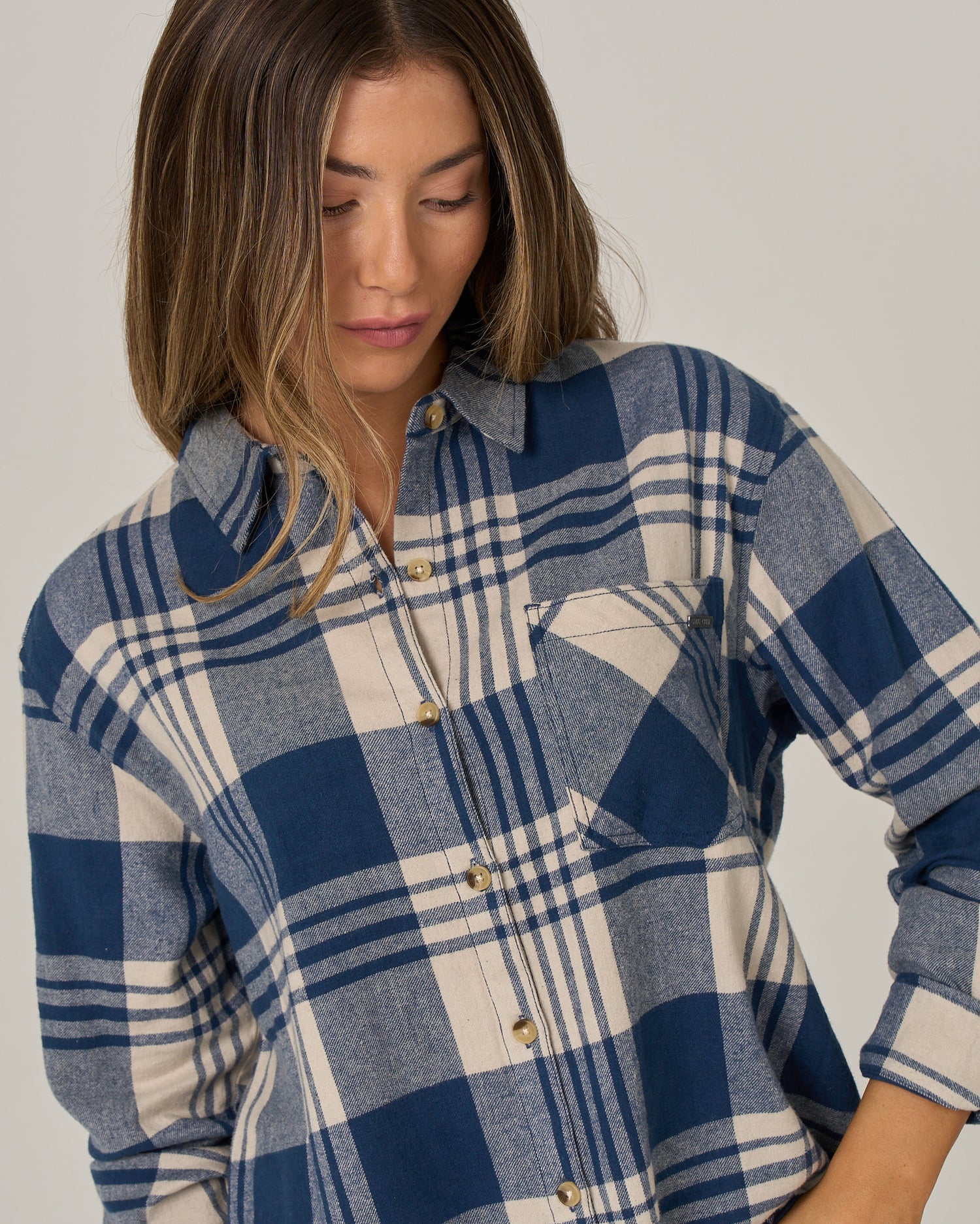 A person with shoulder-length brown hair wears the Salty Crew Out West Flannel - Navy, an oversized blue and beige plaid cotton button-up shirt in a relaxed western style, looking down with a neutral expression against a plain background.