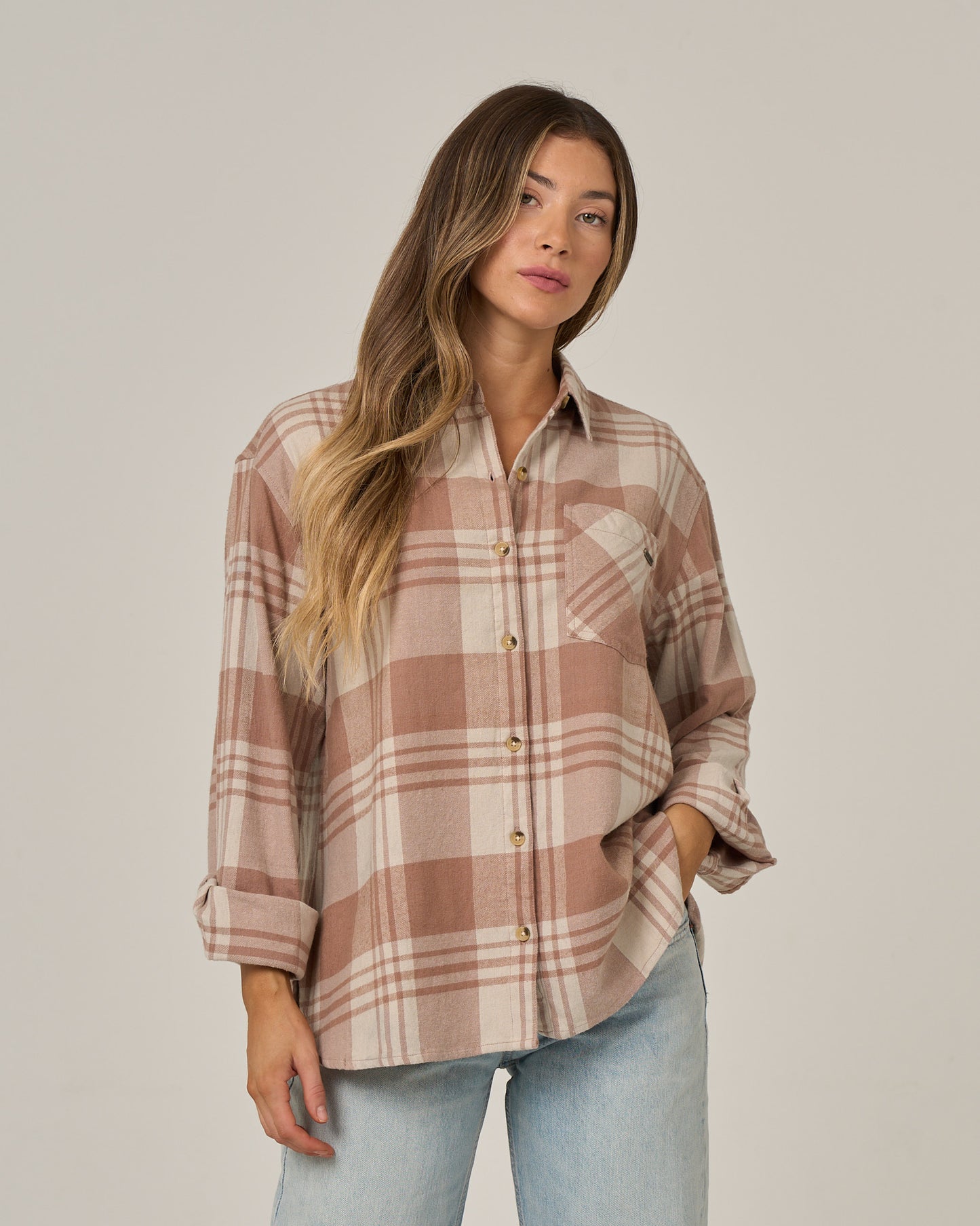 A woman wearing the Salty Crew Out West Flannel in Taupe with rolled sleeves and light blue jeans stands against a plain background, looking at the camera with one hand in her pocket.
