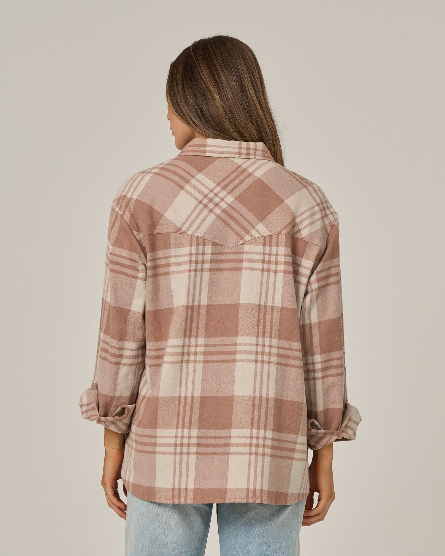 A person with long brown hair, wearing the Salty Crew Out West Flannel in Taupe—an oversized cotton flannel shirt featuring a beige and pink plaid pattern with rolled-up sleeves—paired with light blue jeans, stands facing away from the camera against a plain background.