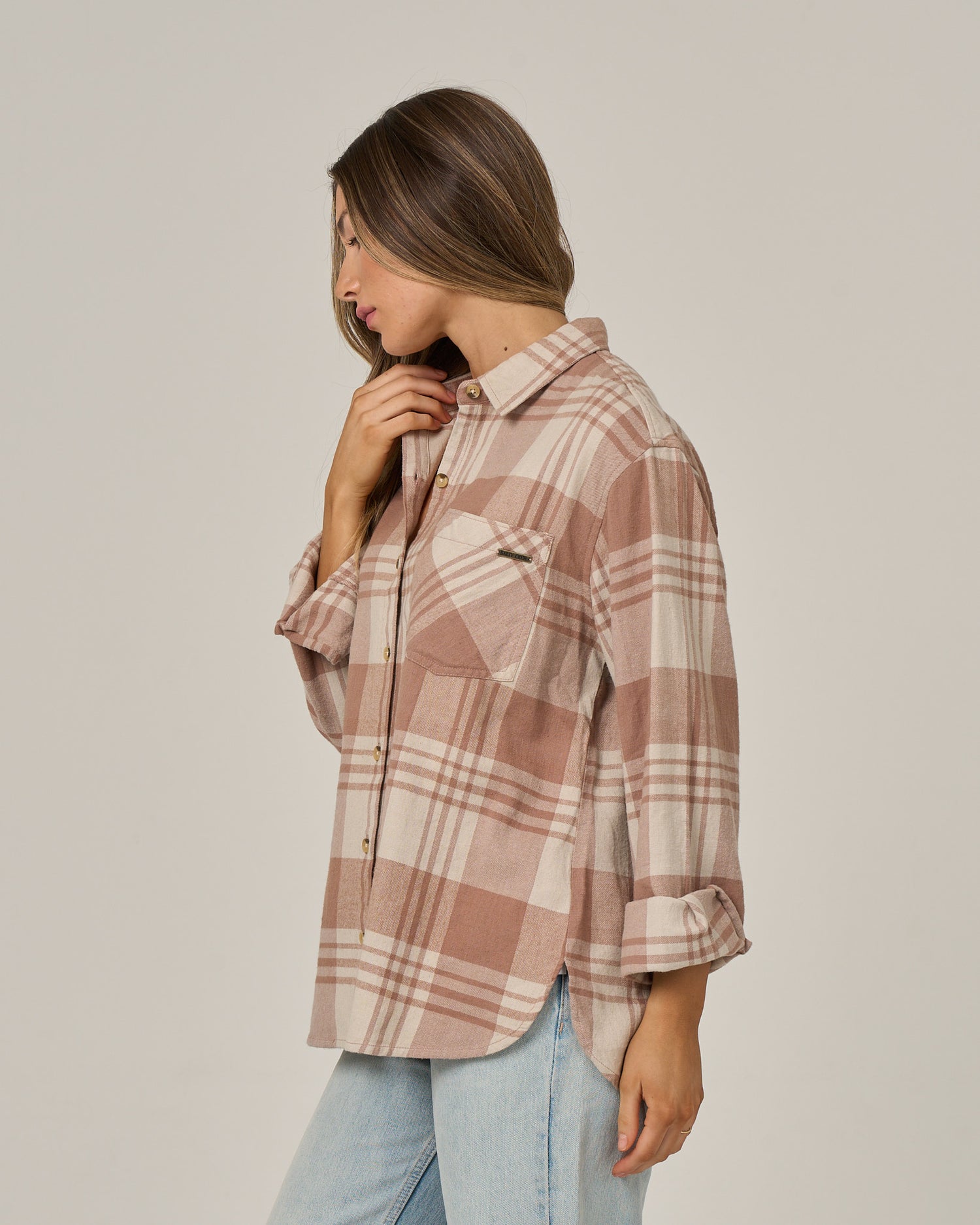 A woman wearing the Salty Crew Out West Flannel in taupe—a western-style, oversized, plaid long-sleeve shirt—paired with light blue jeans, stands sideways touching her neck and looking down against a plain background.