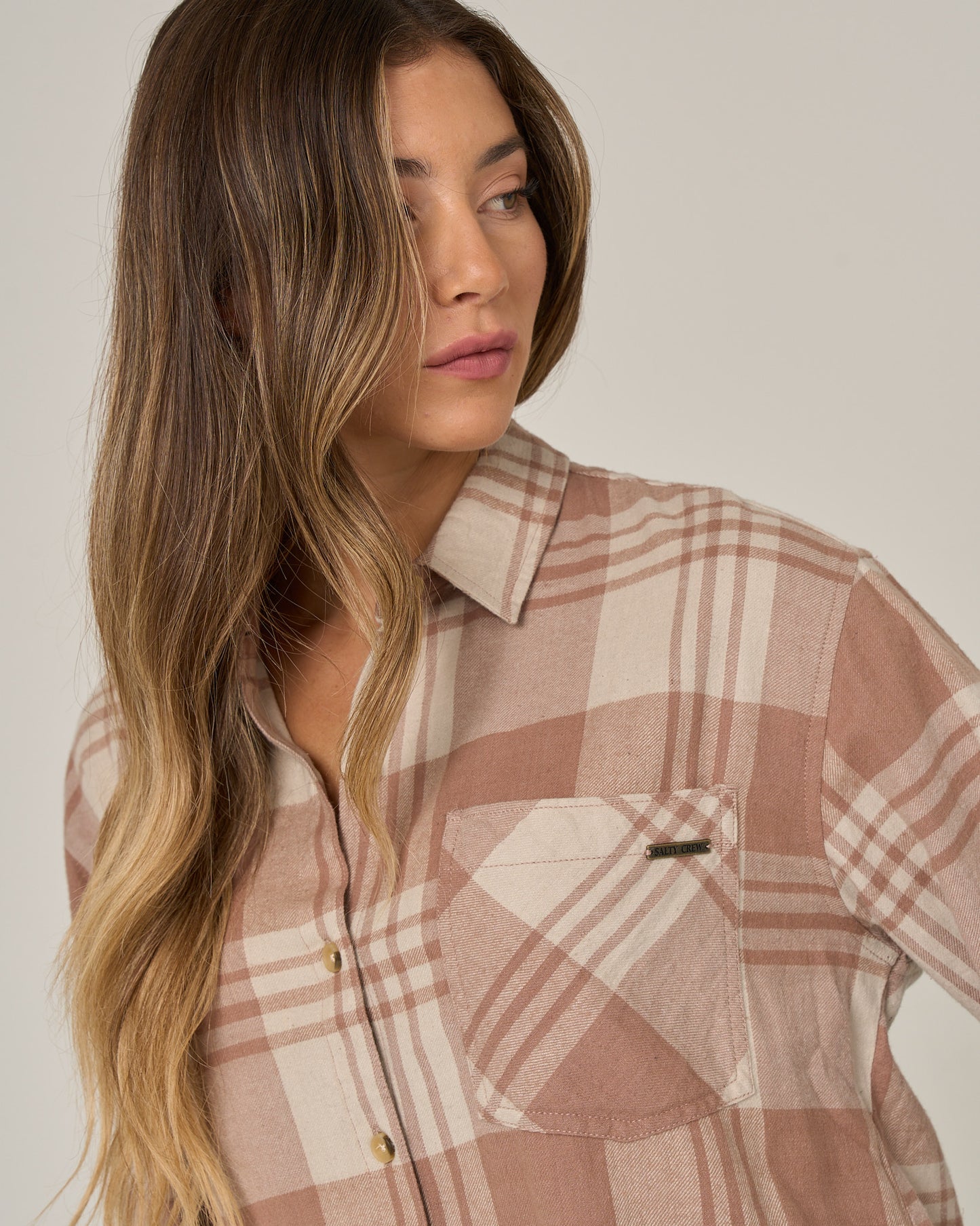 A woman with long wavy hair looks to the side against a plain background, embodying a relaxed western style in the Salty Crew Out West Flannel in Taupe.