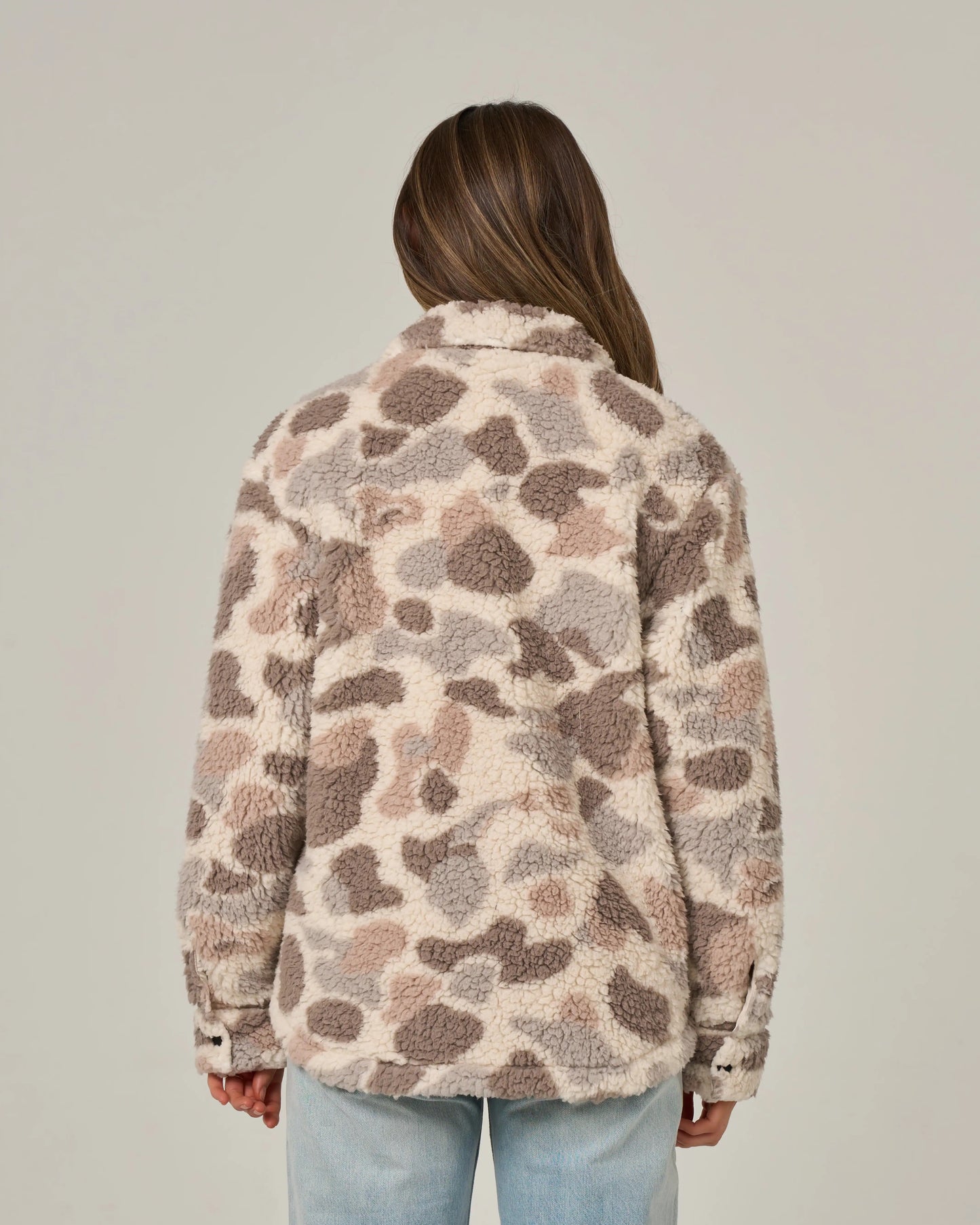 A person with long brown hair, seen from behind, wears the Salty Crew Calm Seas Shacket in Sand Camo—a plush sherpa jacket featuring quilted chest pockets and a fuzzy, spotted beige and brown pattern—paired with light blue jeans against a plain background.