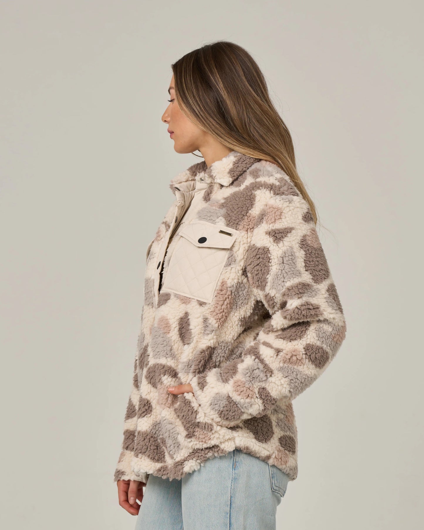 A woman stands in profile wearing the Salty Crew Calm Seas Shacket in Sand Camo, featuring plush sherpa fabric with quilted chest pockets, paired with light blue jeans against a plain background.