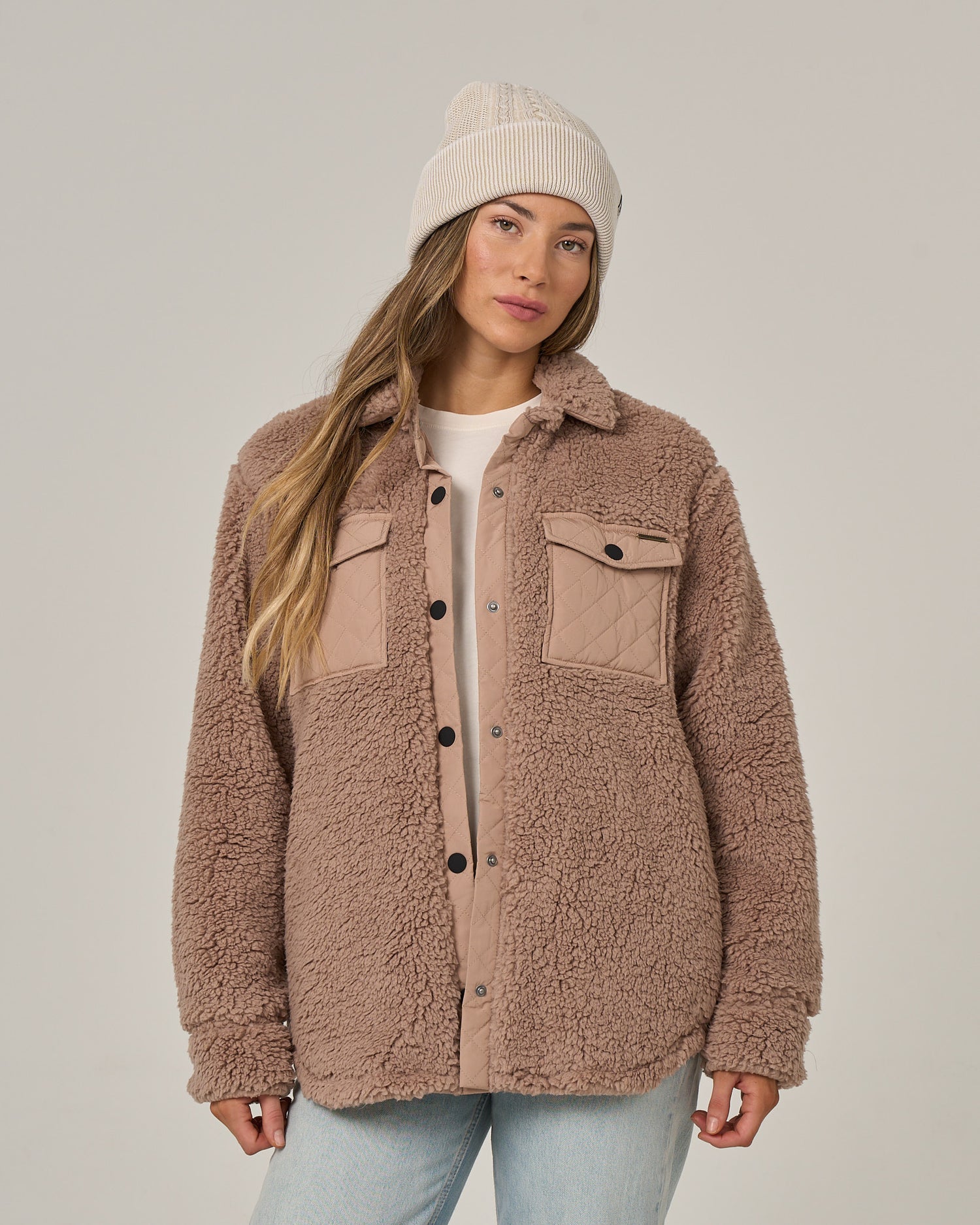 A person wearing a beige knit beanie, a white shirt, light blue jeans, and the Salty Crew Calm Seas Shacket in taupe—with plush sherpa fabric, quilted chest pockets, and snap front closure—stands against a plain light background.
