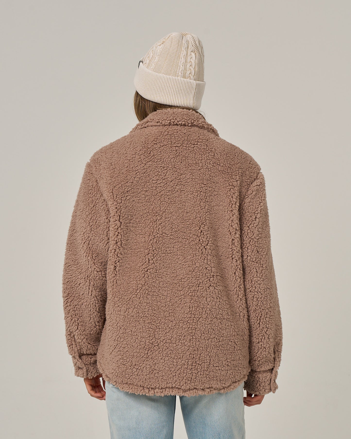 A person facing away, wearing the Salty Crew Calm Seas Shacket in taupe—a plush sherpa jacket with quilted chest pockets—paired with a beige knit beanie and light blue jeans, set against a plain off-white background.