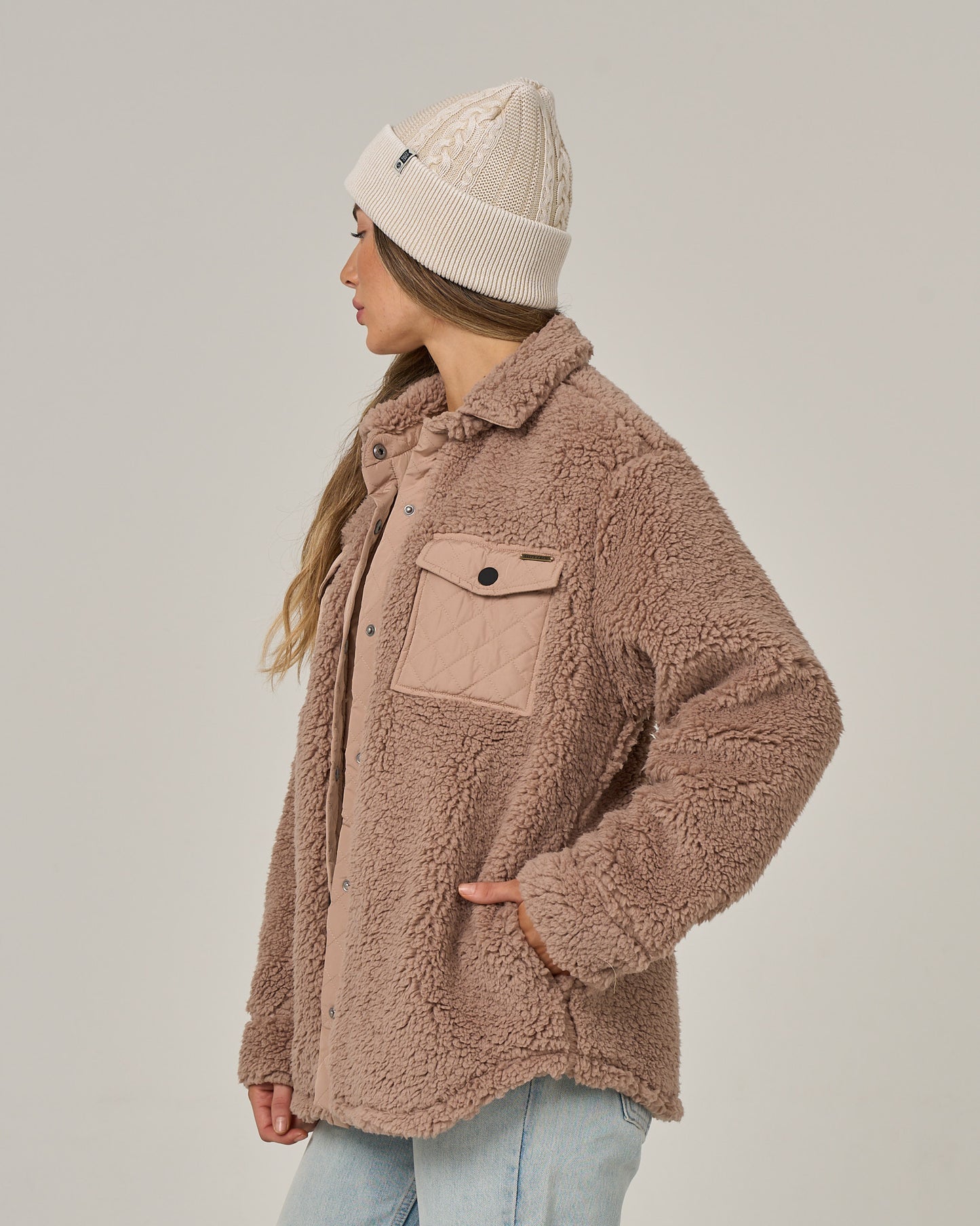 A woman stands sideways against a plain light background, looking left. She wears the Salty Crew Calm Seas Shacket in taupe—a plush sherpa jacket featuring a snap front closure—paired with light blue jeans and a white knit beanie. Her long hair falls over her shoulder as she rests one hand on her hip.