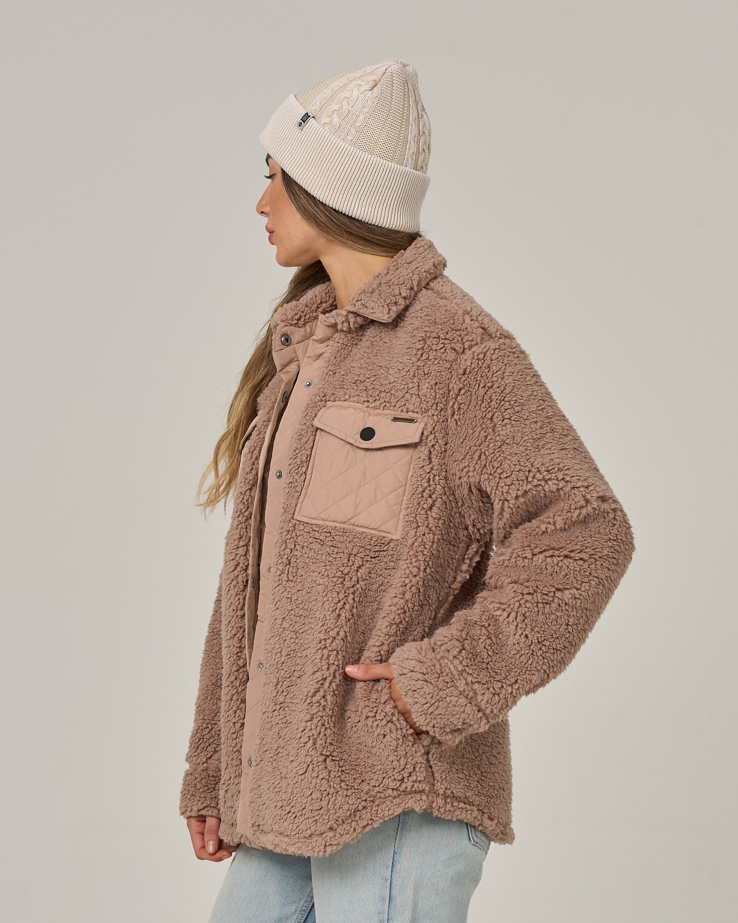 A woman stands sideways against a plain light background, looking left. She wears the Salty Crew Calm Seas Shacket in taupe—a plush sherpa jacket featuring a snap front closure—paired with light blue jeans and a white knit beanie. Her long hair falls over her shoulder as she rests one hand on her hip.