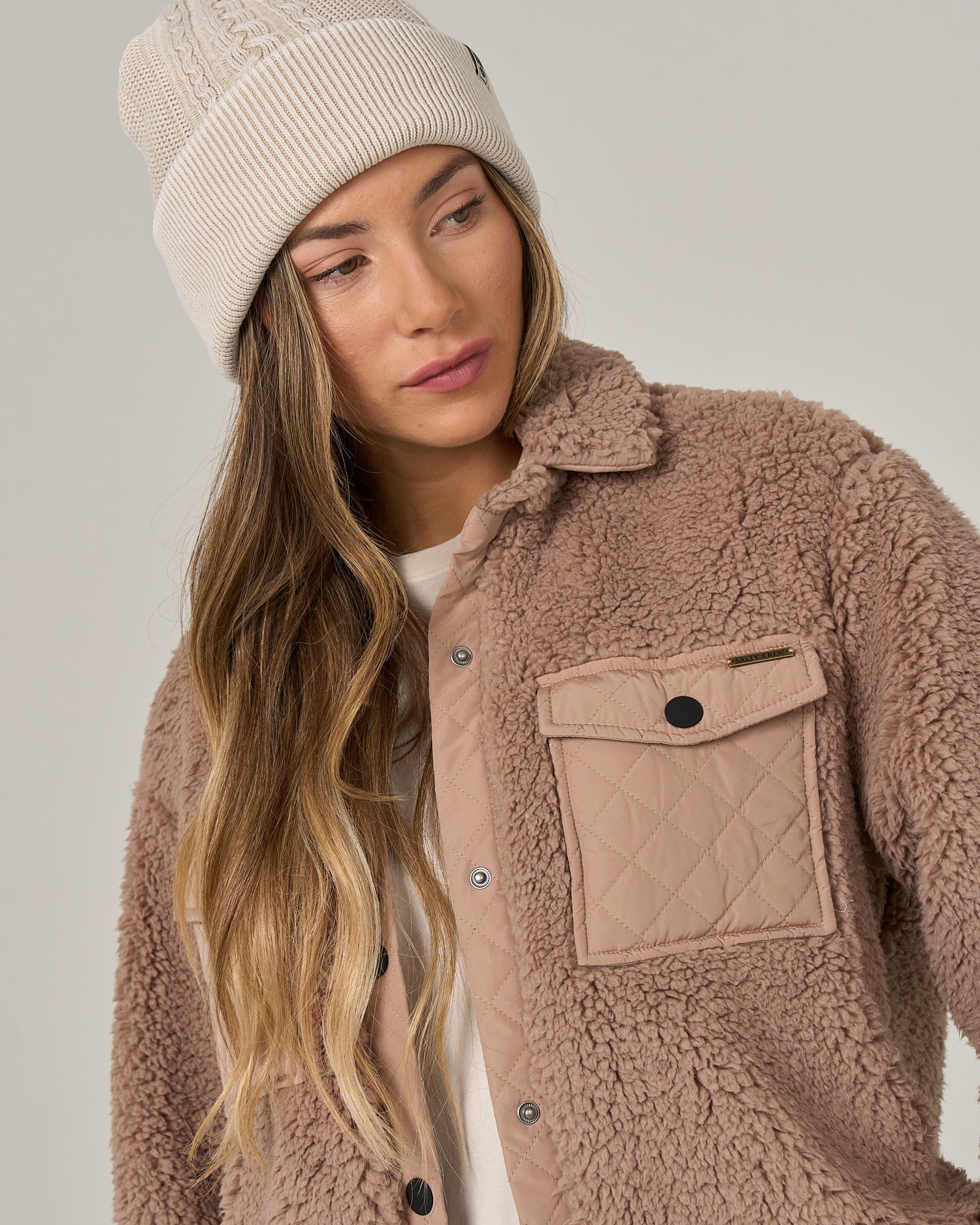 A woman with long, wavy hair wears the Salty Crew Calm Seas Shacket in taupe—a plush sherpa jacket with a snap front closure and quilted pocket—paired with a cream knit beanie, as she looks slightly down and to the side.