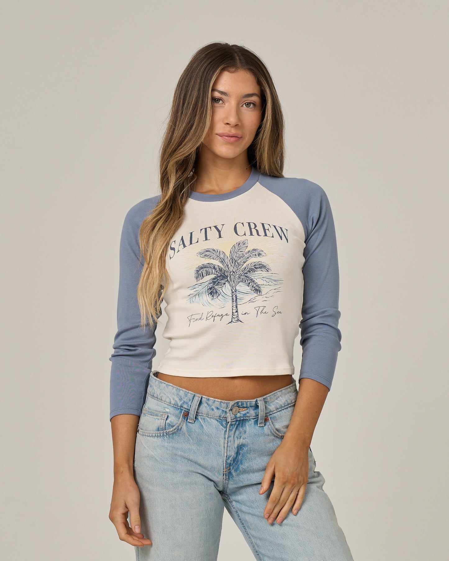A woman stands facing forward against a neutral background, one hand in her pocket, wearing the Salty Crew Pretty Palms Long Sleeve tee in Flint with a palm tree screen print.