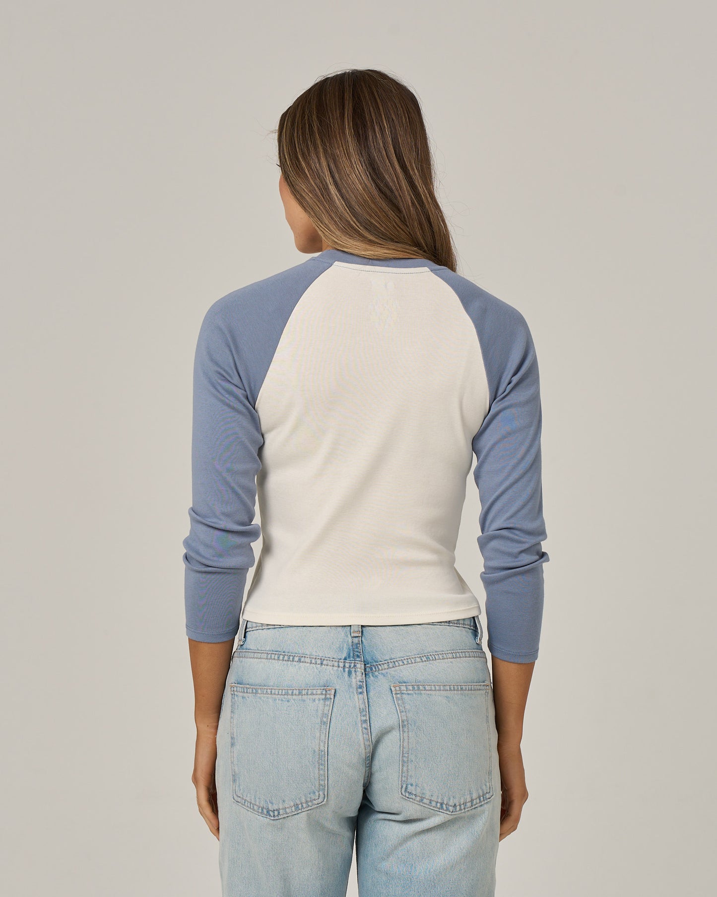A person with long brown hair, wearing the Salty Crew Pretty Palms Long Sleeve - Flint styled as a raglan tee and light blue jeans, stands facing away from the camera against a plain background.
