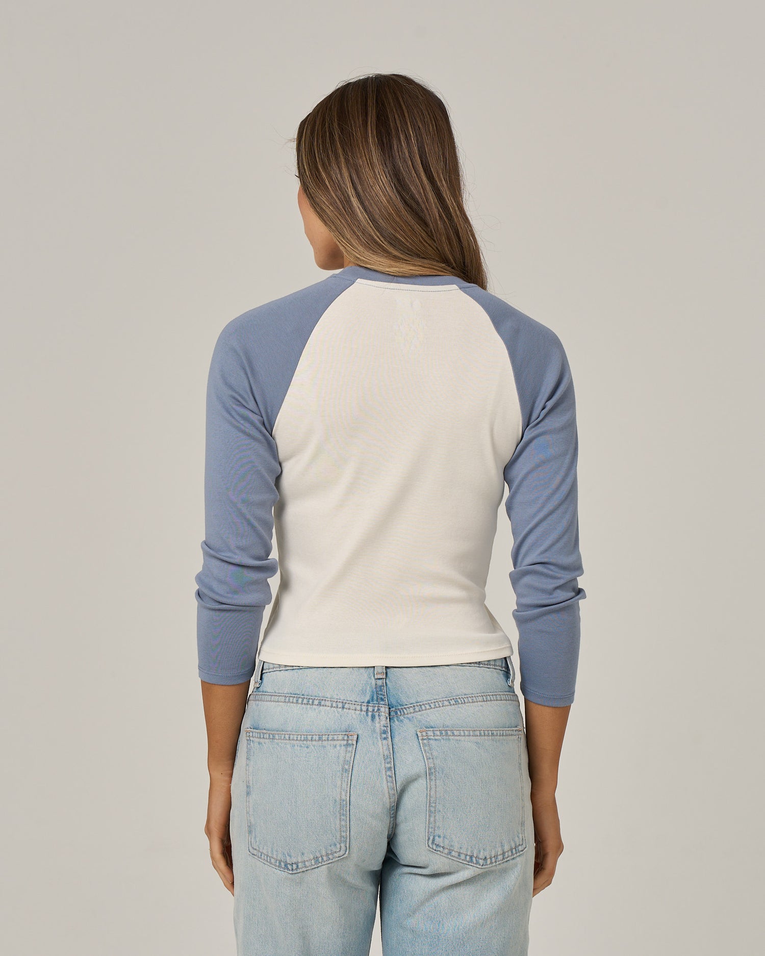 A person with long brown hair, wearing the Salty Crew Pretty Palms Long Sleeve - Flint styled as a raglan tee and light blue jeans, stands facing away from the camera against a plain background.